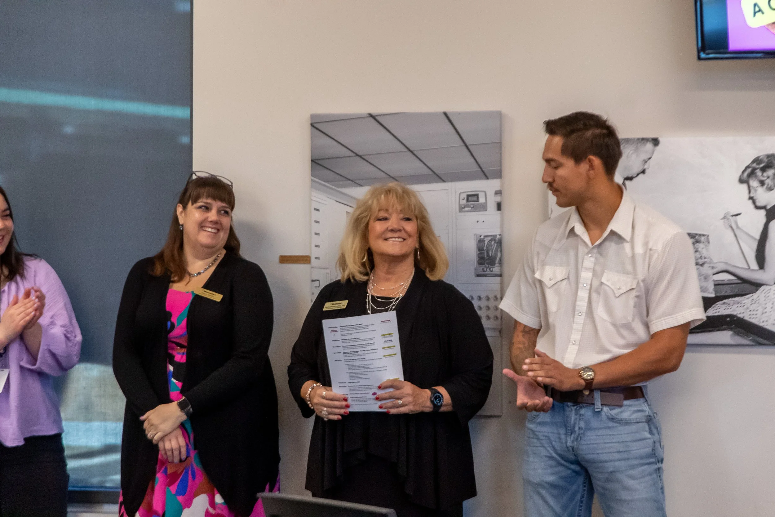 Group of four people standing indoors, three women and one man, smiling and engaged in conversation. One woman is holding a sheet of paper, and they appear to be at a professional or social event.