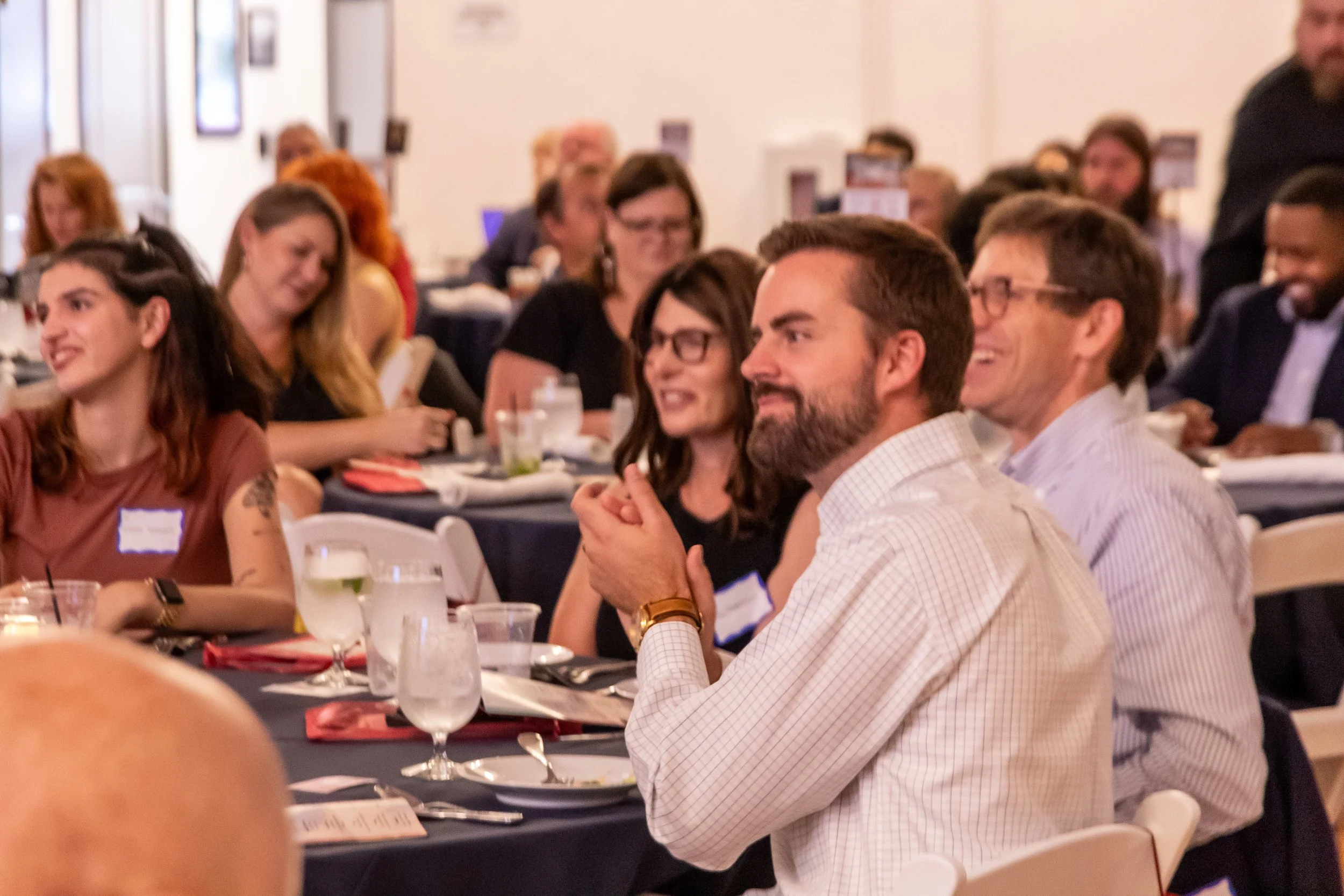 People sitting at tables in a banquet or conference setting, listening attentively and smiling.