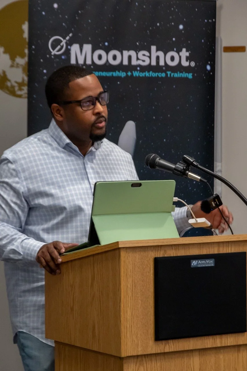 A man giving a presentation at a podium with a microphone in front of a banner that reads 'Moonshot entrepreneurship + Workforce Training'.