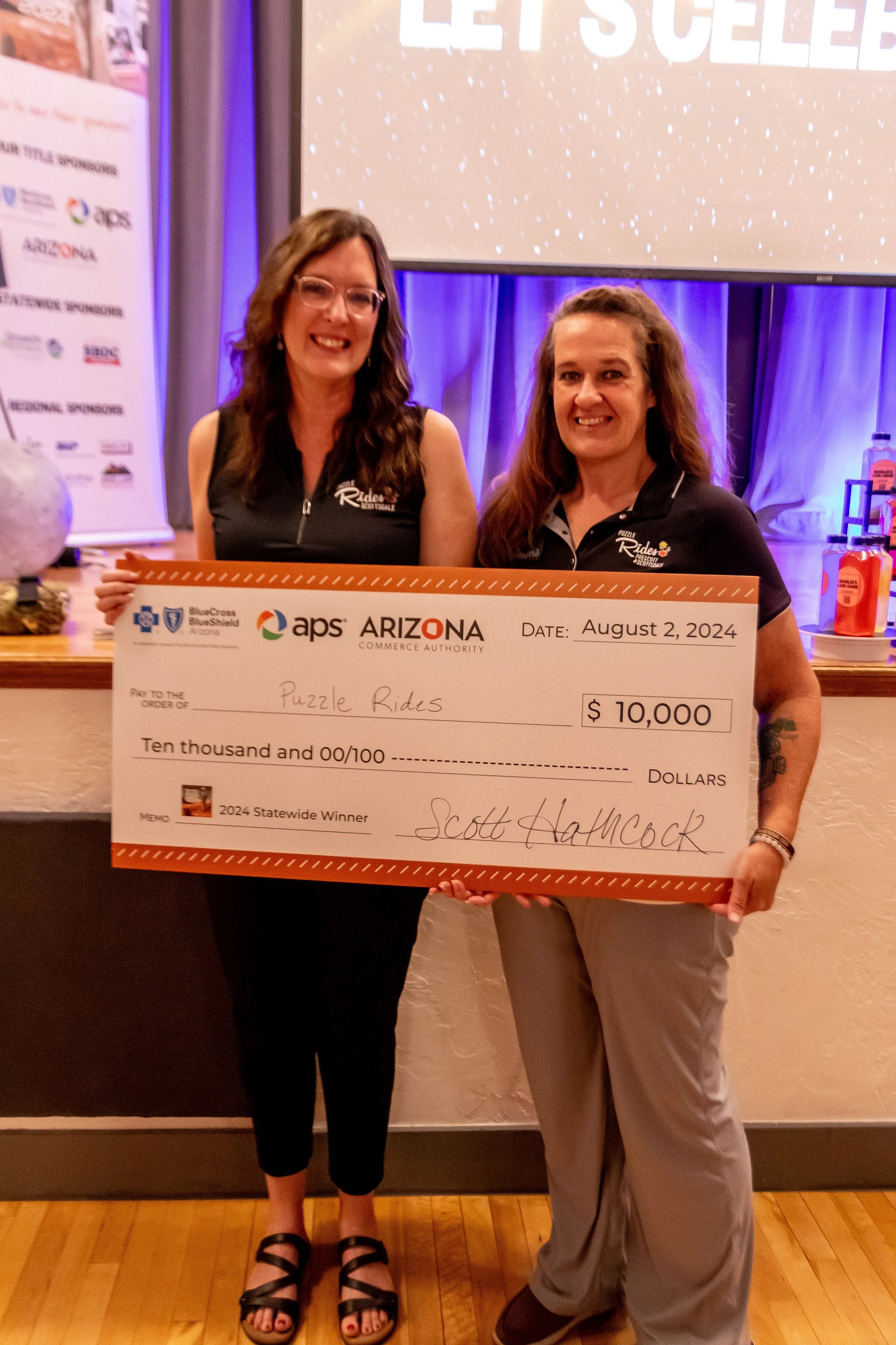 Two women smiling and holding a large check for ten thousand dollars, awarded to Puzzle Rides, at an event in Arizona on August 2, 2024.