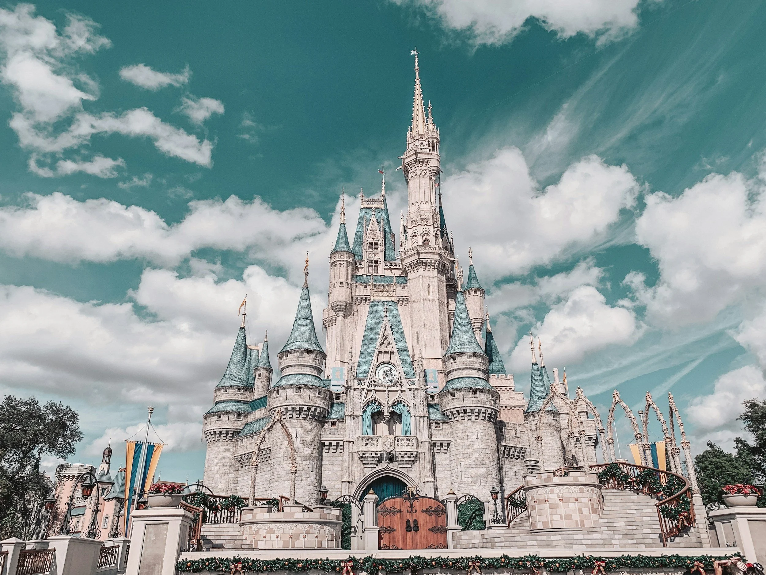 Disney castle with blue turrets and flags, surrounded by a decorated fence, under a partly cloudy sky.