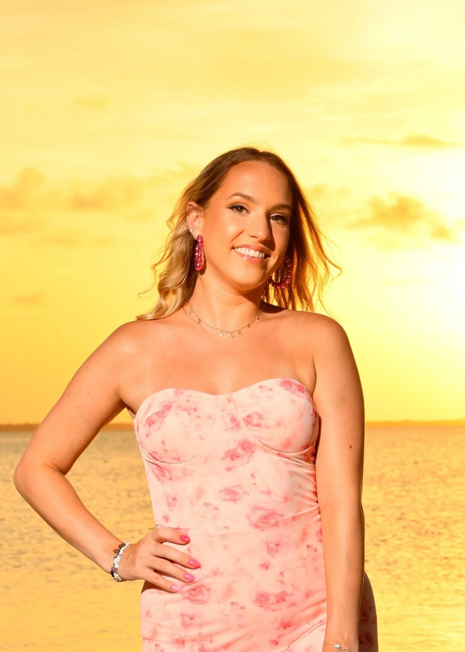 A woman with blonde hair wearing a pink tie-dye strapless dress and pink earrings standing in front of a sunset over water.