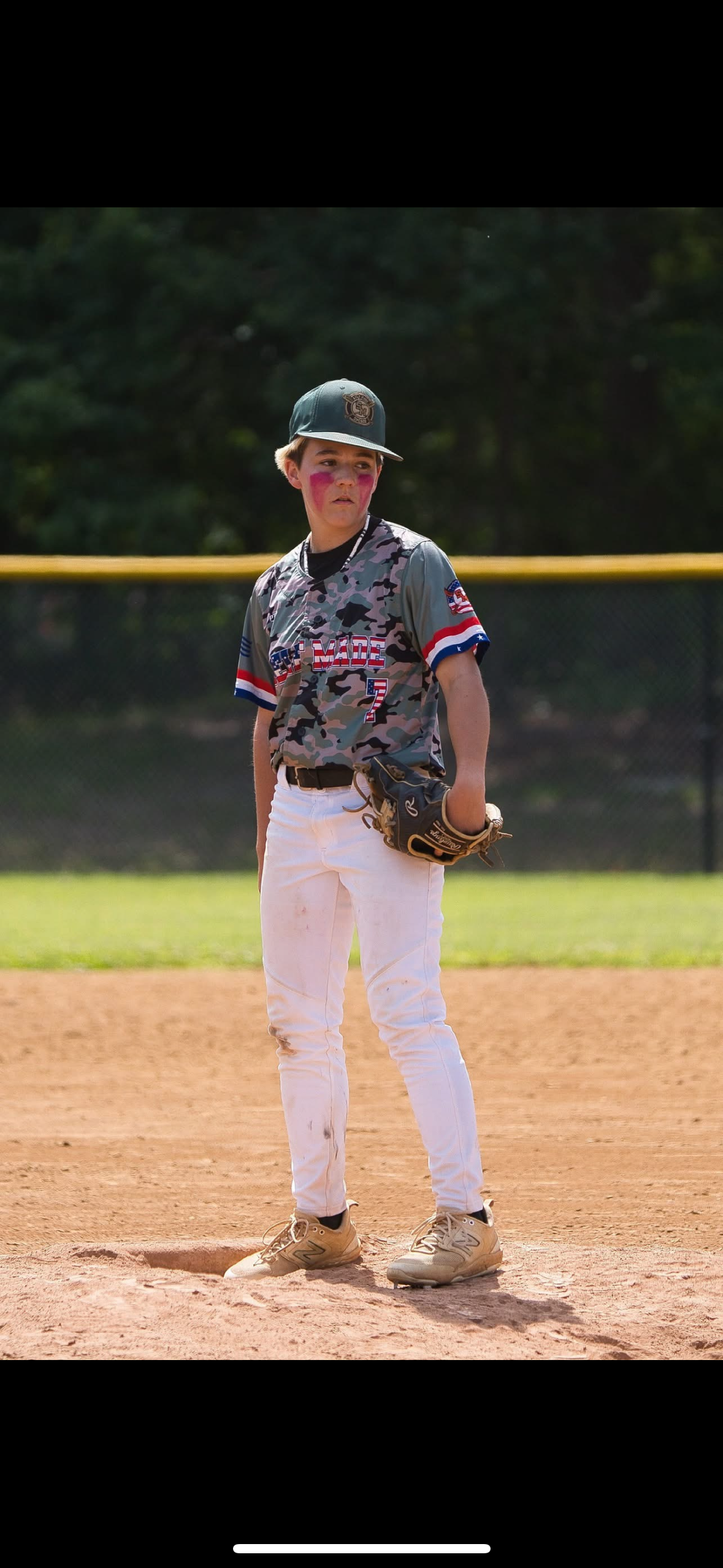 A young baseball player in a camouflage jersey, white pants, and a helmet is preparing to hit a baseball at a baseball field.