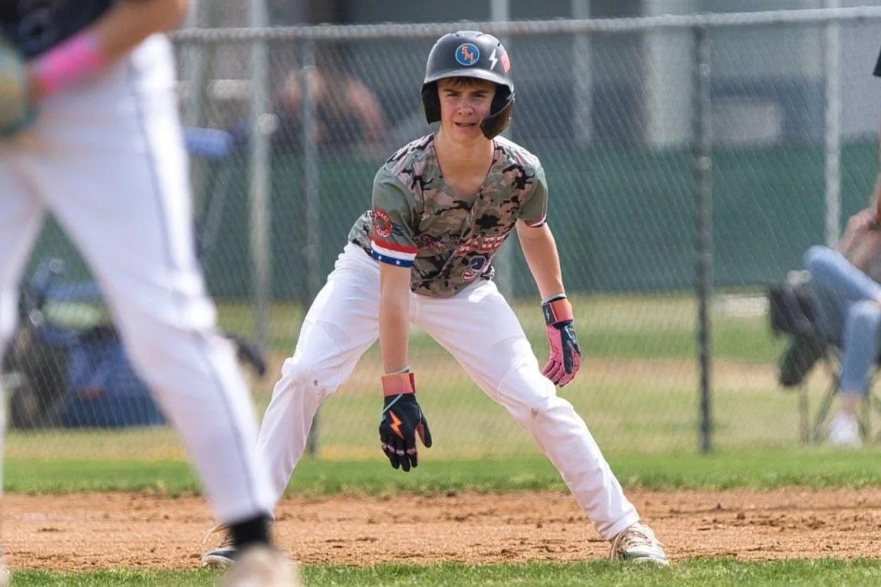 A young baseball player wearing a helmet, camouflage shirt, white pants, and pink batting gloves standing on a baseball field in a ready stance.