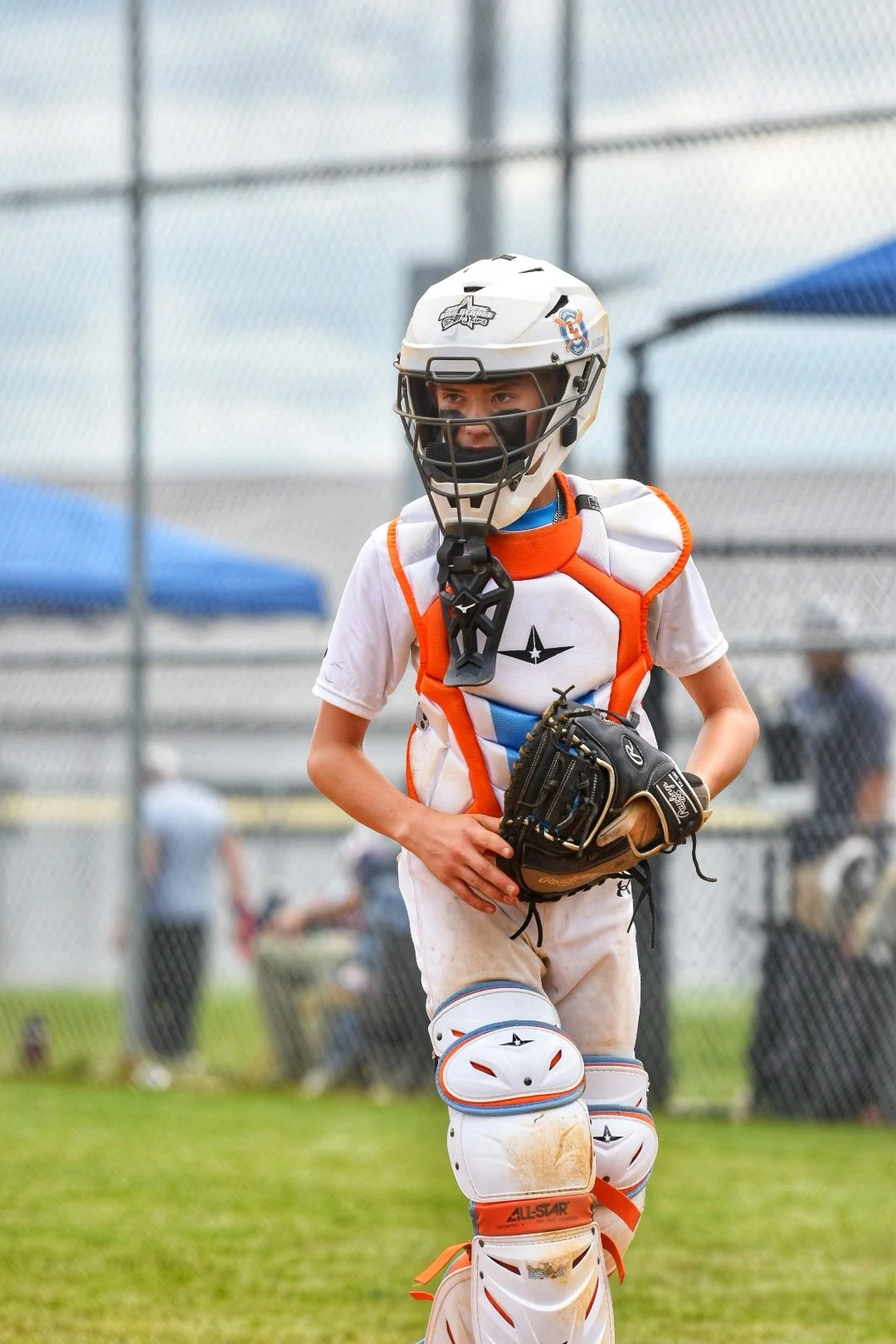A young boy wearing a sports uniform, holding a catcher's helmet on his head and a baseball glove, standing outdoors on a baseball field.