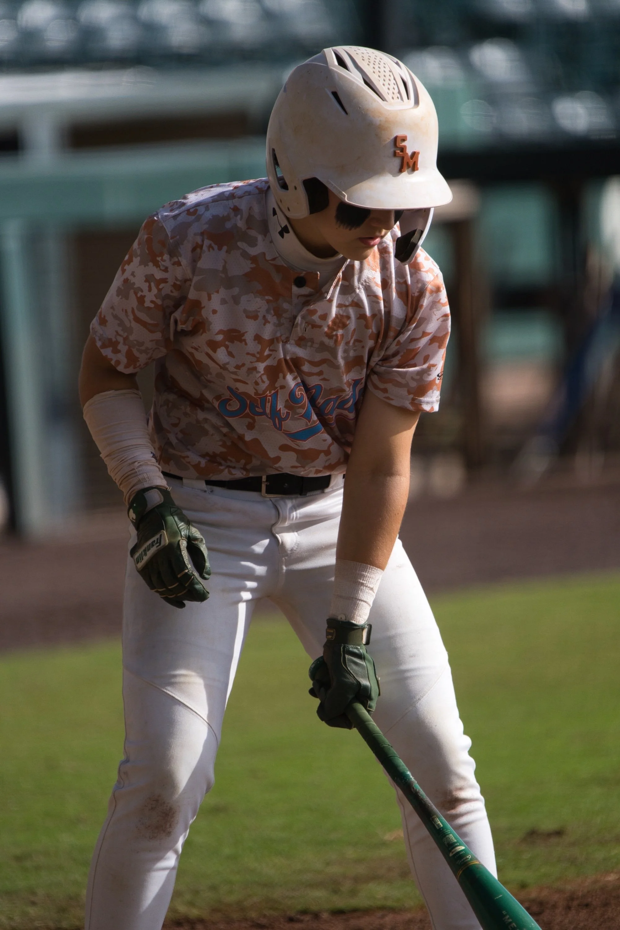 A young baseball player in a camouflage jersey, white pants, and a helmet is preparing to hit a baseball at a baseball field.