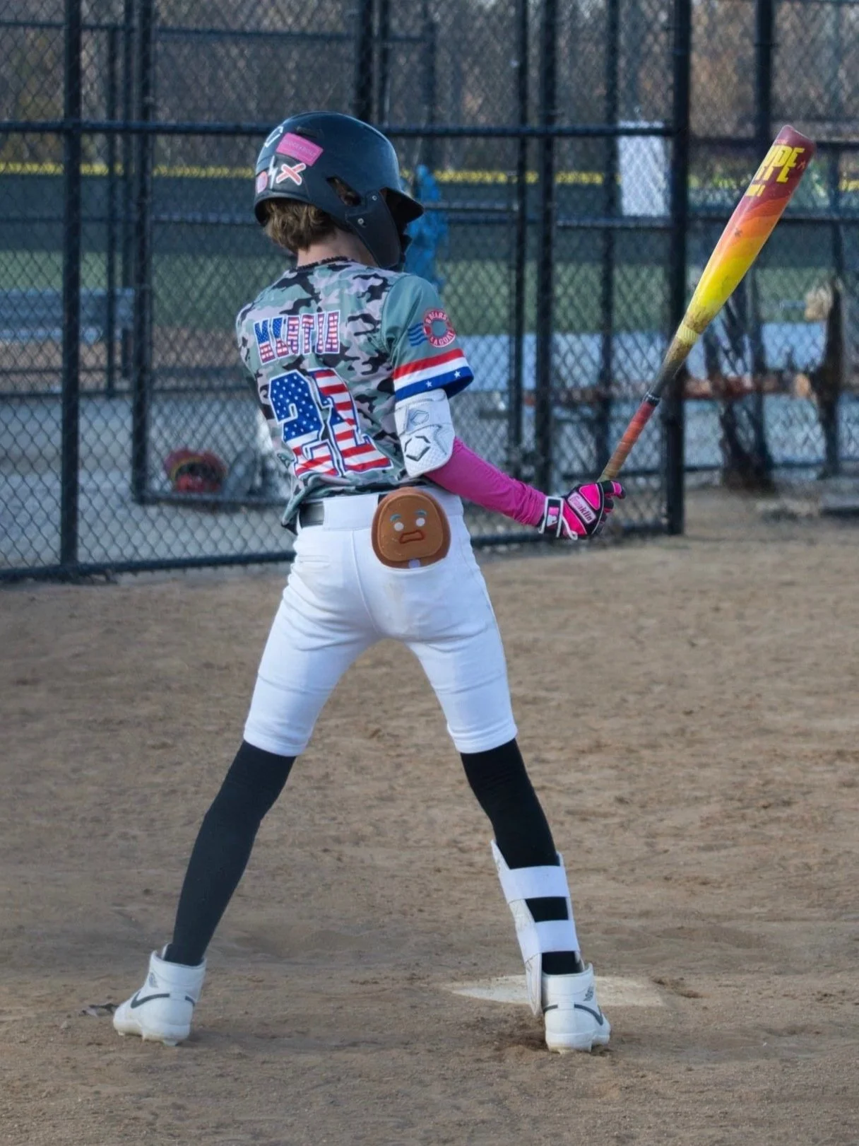 A young baseball player in a camouflage jersey and black kneepad is pitching a ball on the field. He is wearing a green cap and pink arm sleeves, with a glove on his left hand. He is mid-throw on a dirt infield, with an outfield fence and trees in the background.