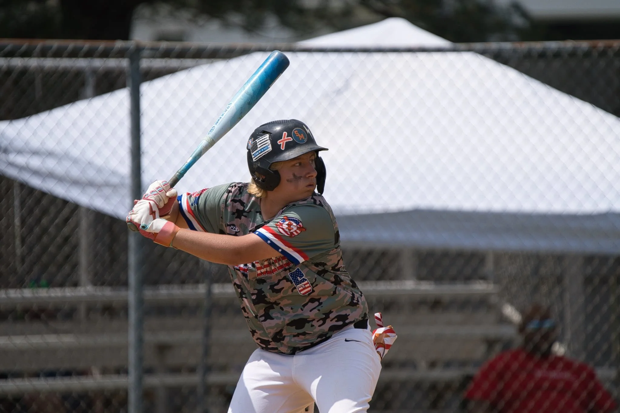 A baseball player in camouflage uniform and a black helmet preparing to swing a bat at a pitch during a game.