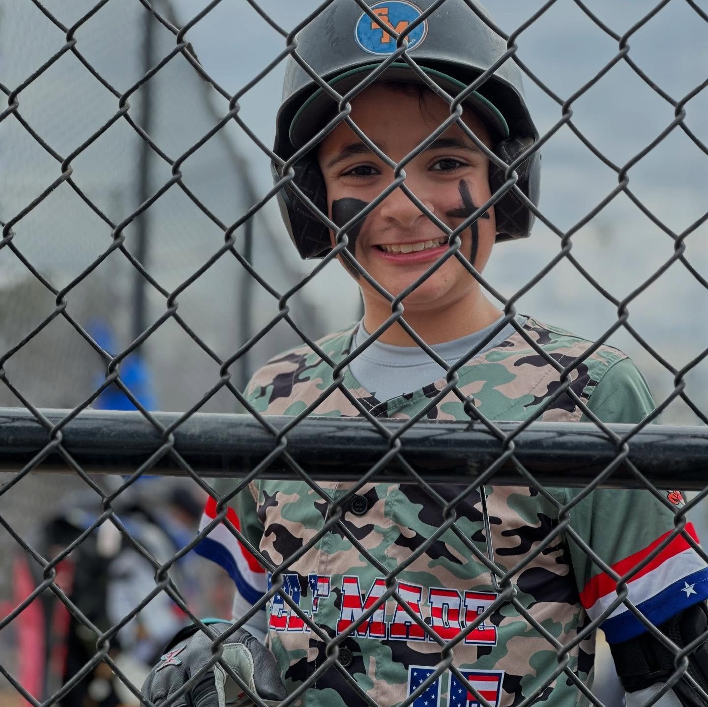 A young boy smiling behind a chain-link fence, wearing a football helmet and camouflage uniform, with face paint.