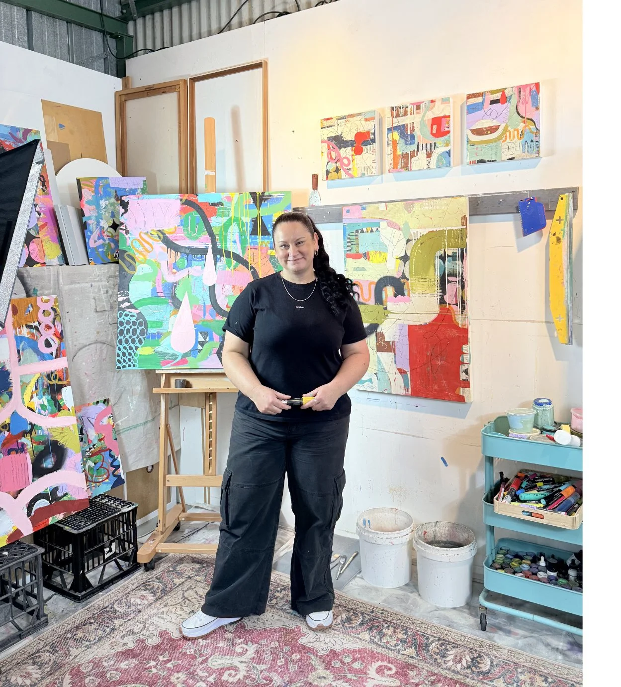 The artist Angelea Van Dam stands in front of colorful abstract paintings in her Salamander Bay NSW Australia art studio.