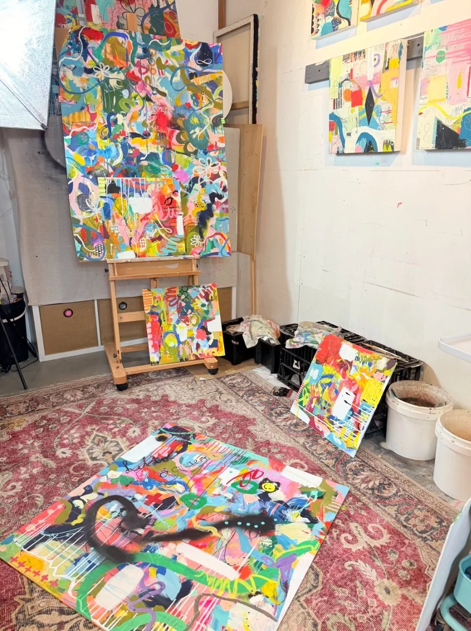 Art studio with abstract paintings on the wall, on the floor, and a canvas on an easel; various paint supplies and buckets on the floor.