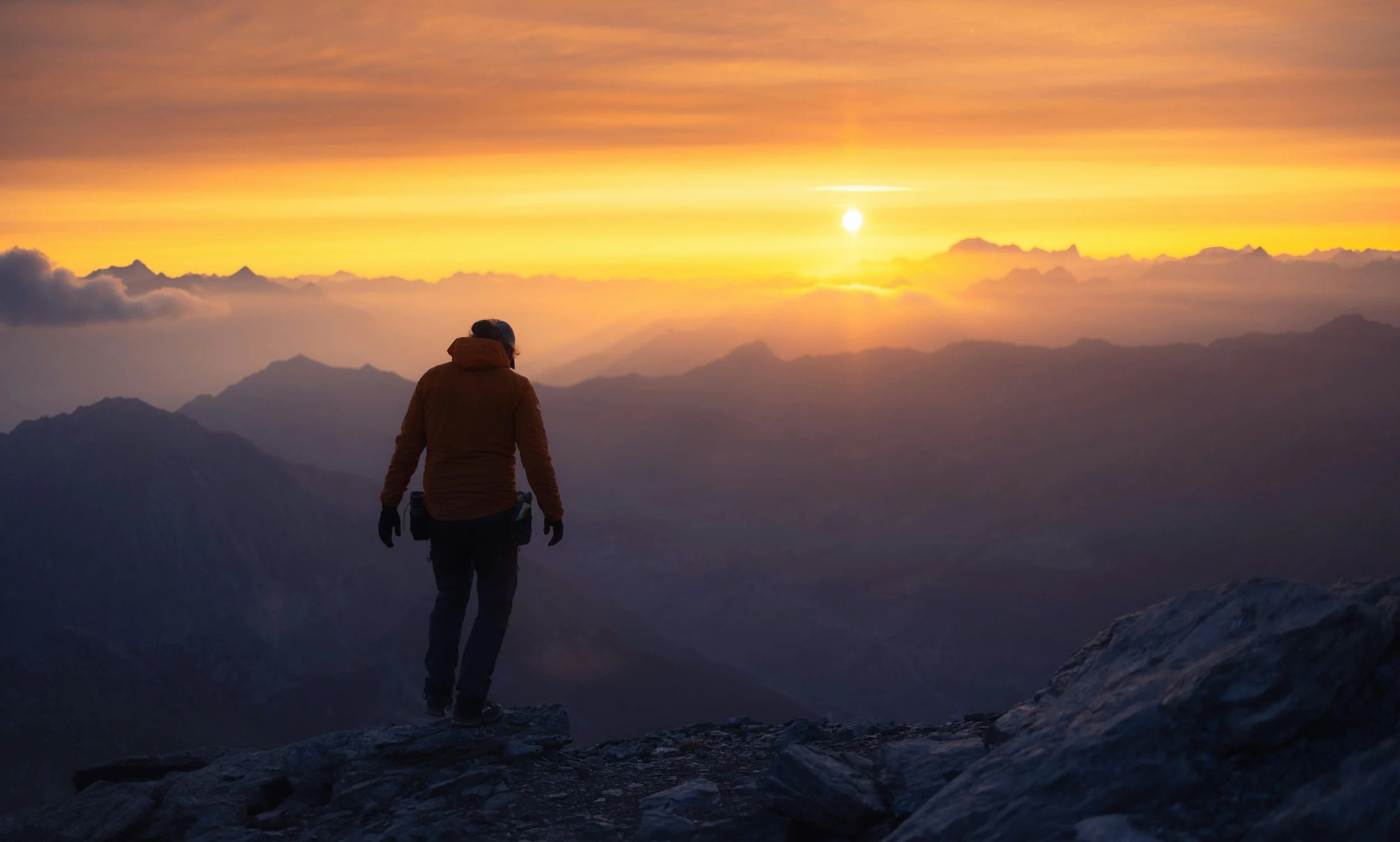 Person standing on a rocky mountain ledge during sunset, overlooking a range of mountains with orange and yellow sky.