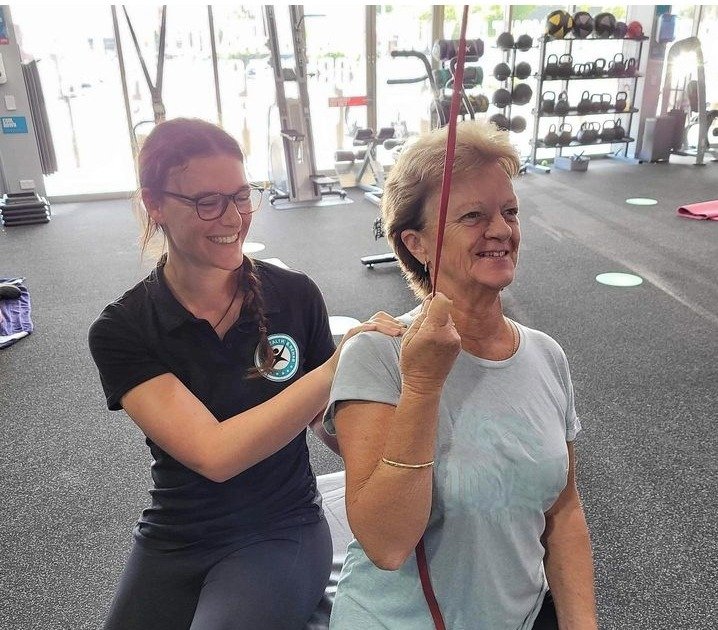 A woman is holding a resistance band above her head while another woman exercises with her in a gym.
