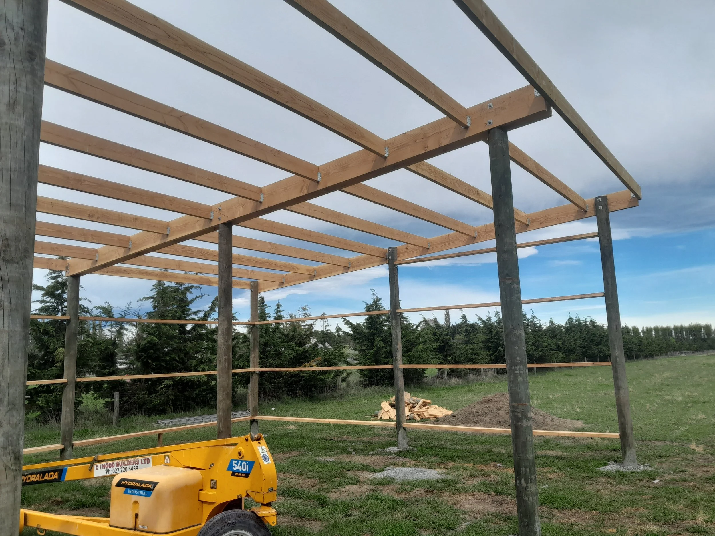 Construction site with wooden framing and support poles for building a structure, with a yellow construction machine in the foreground and a grassy field with trees in the background.