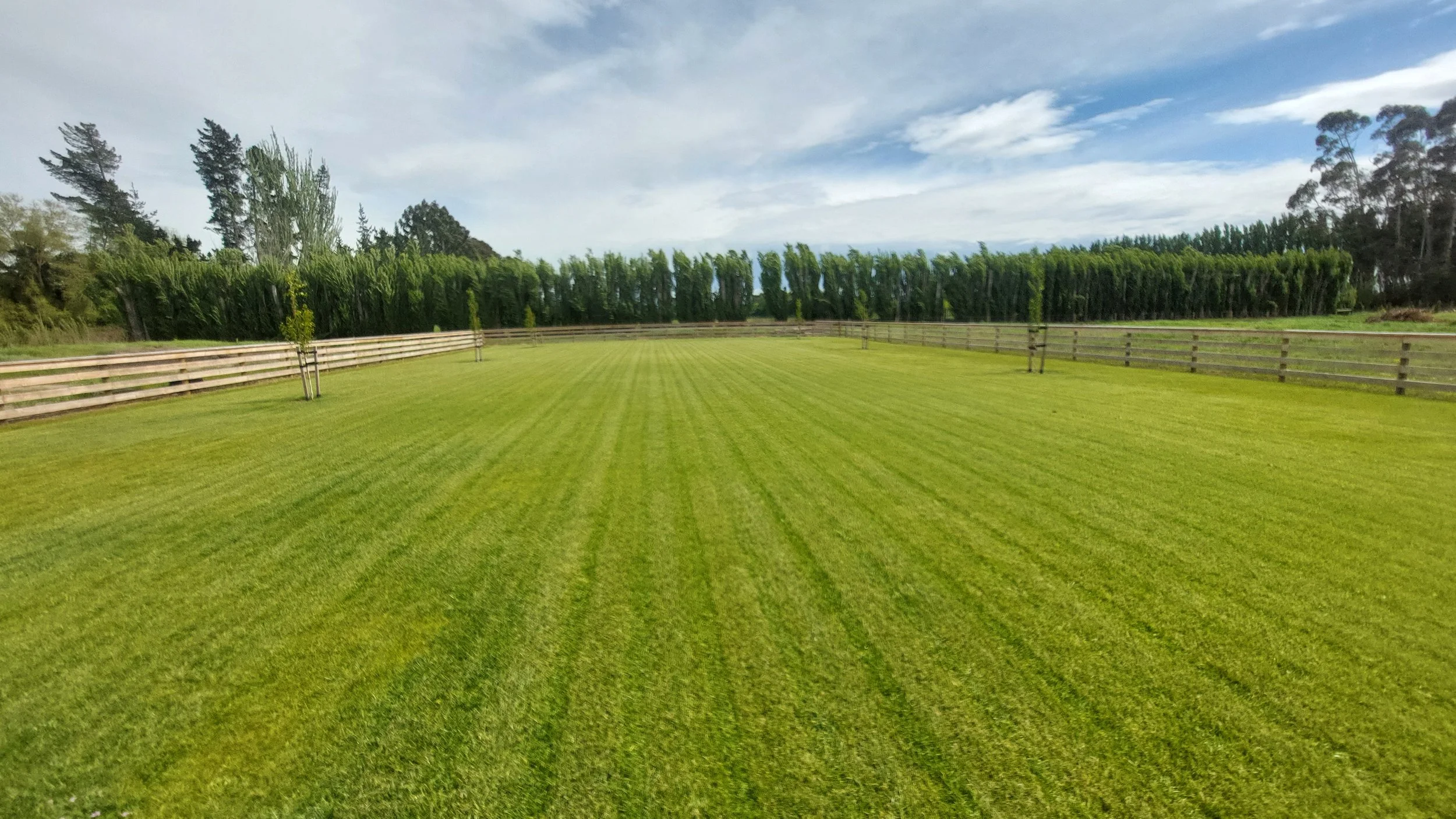 A well-maintained, green grassy field with a wooden fence on three sides, trees in the background, and a partly cloudy sky overhead.