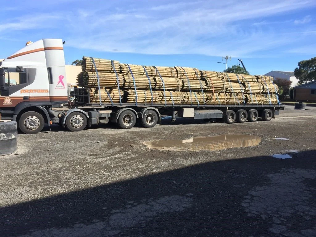 A semi-truck with a flatbed trailer carrying multiple bundles of wooden poles or logs, parked on a gravel lot with a small puddle and a shadow in the foreground, under a partly cloudy blue sky.