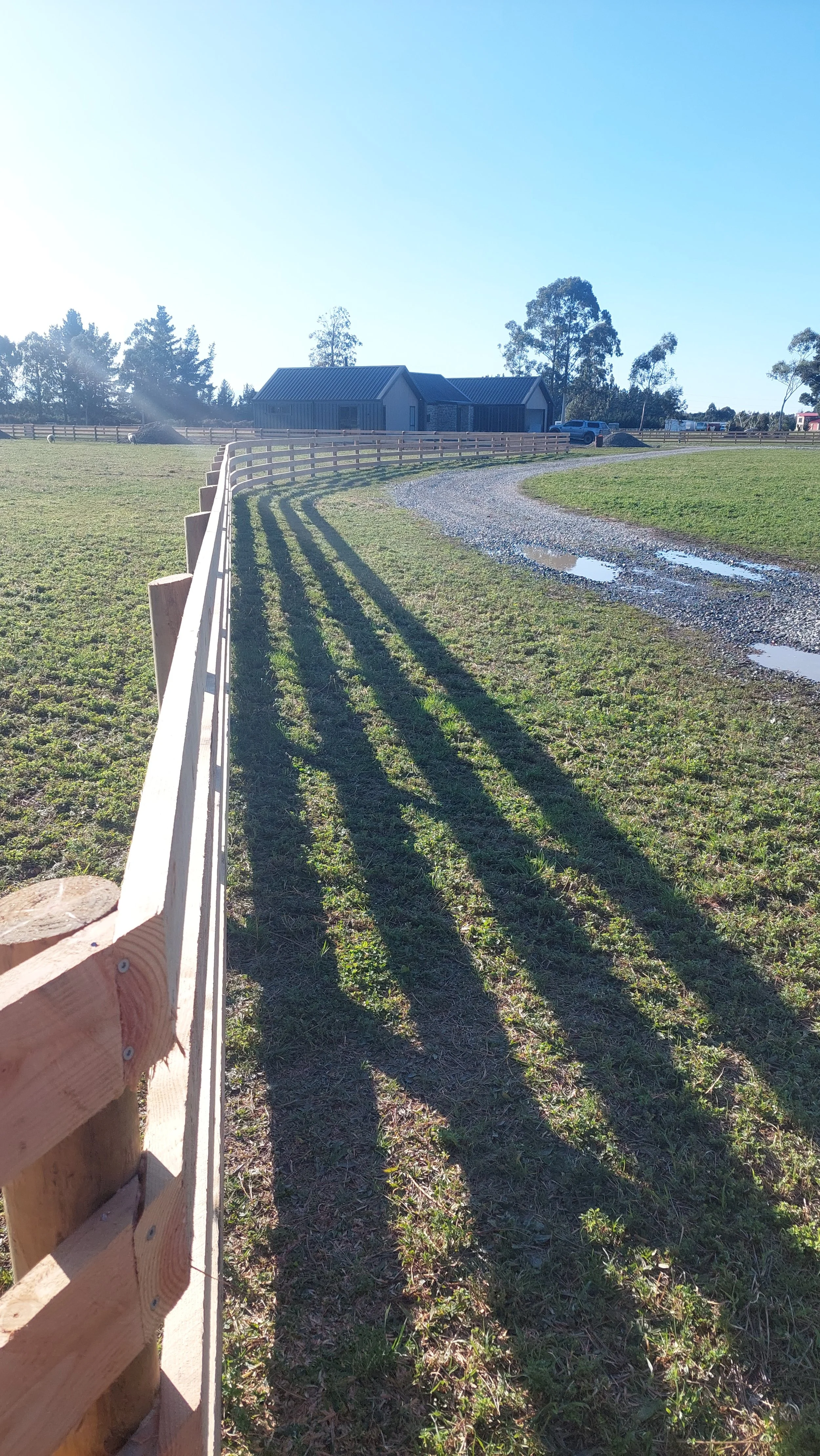 Sunlit rural scene with a gravel driveway, a wooden fence casting long shadows, a green grassy field, and a barn or house with a blue metal roof in the distance.