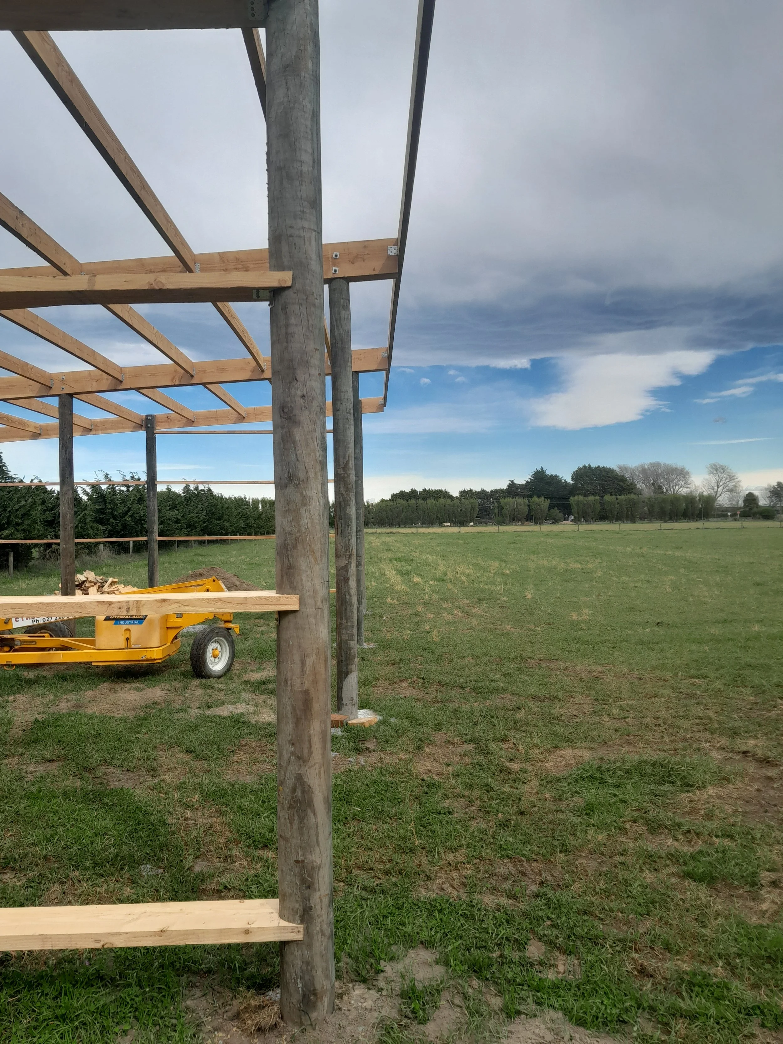 Construction of a wooden structure outdoors on grassy land with a cloudy sky in the background.