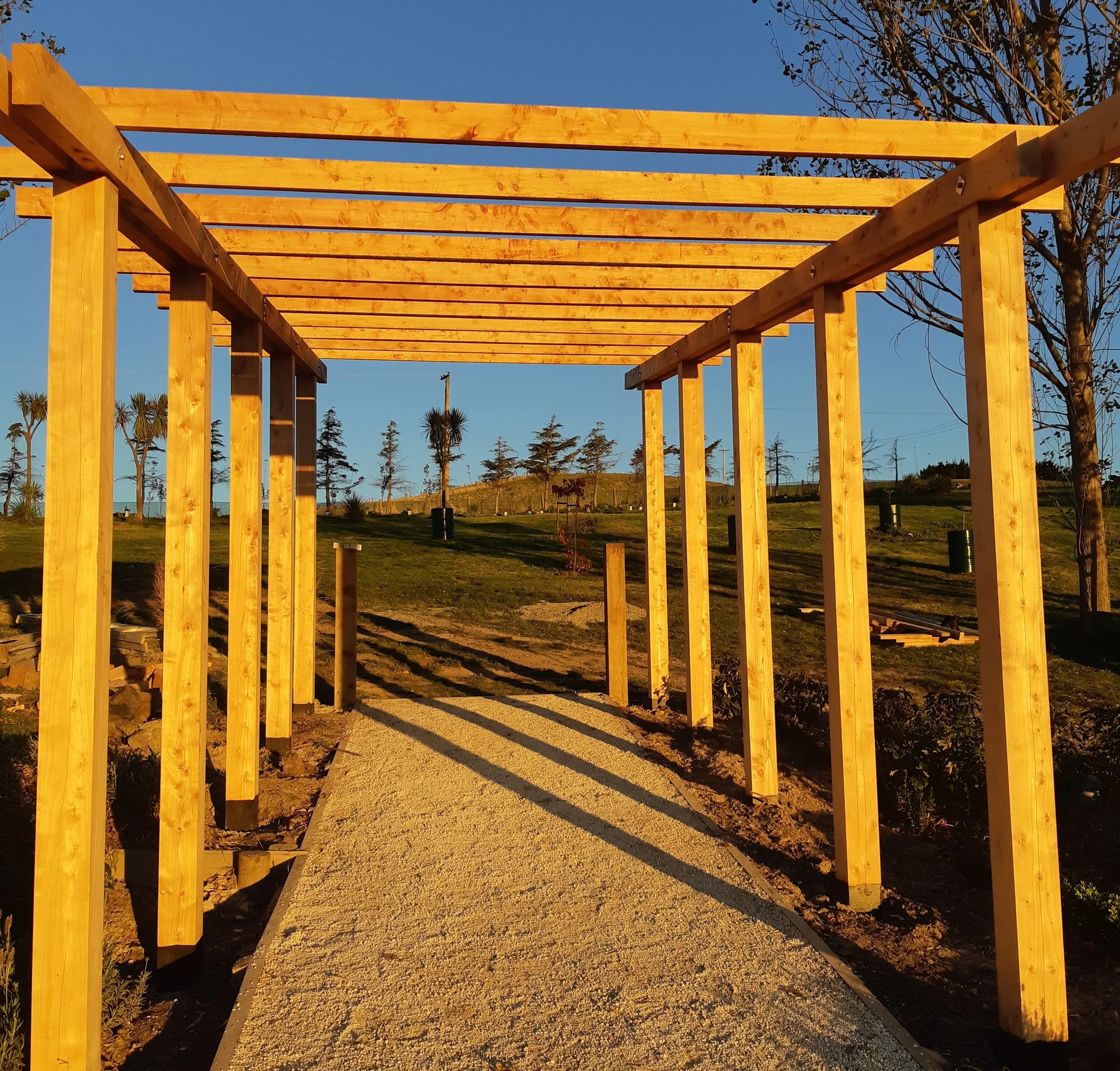 Wooden pergola under construction over a gravel pathway in a park with trees and a clear blue sky in the background.