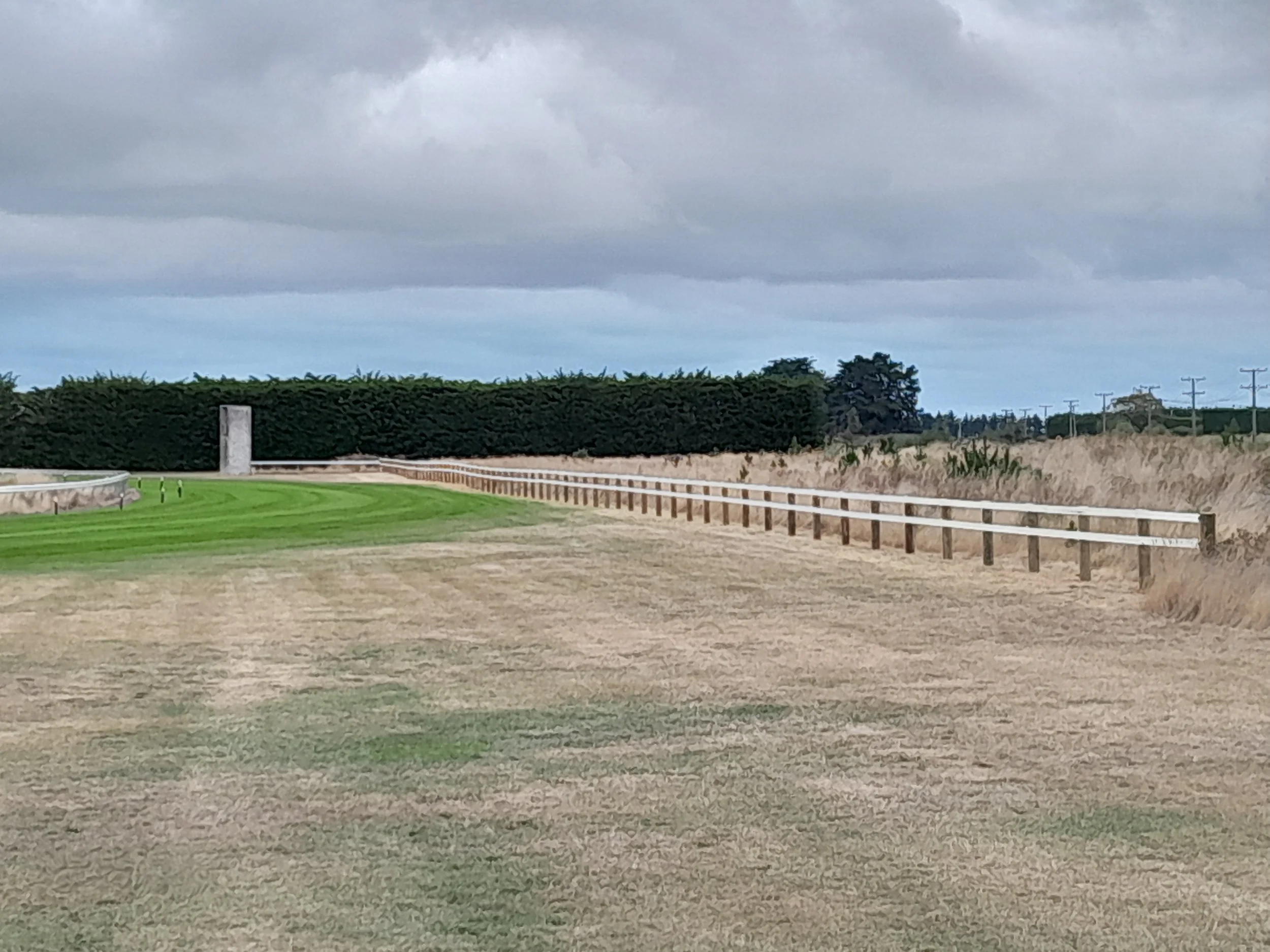 A golf course with a green and sand trap, a wooden fence, and a line of trees in the background under a cloudy sky.