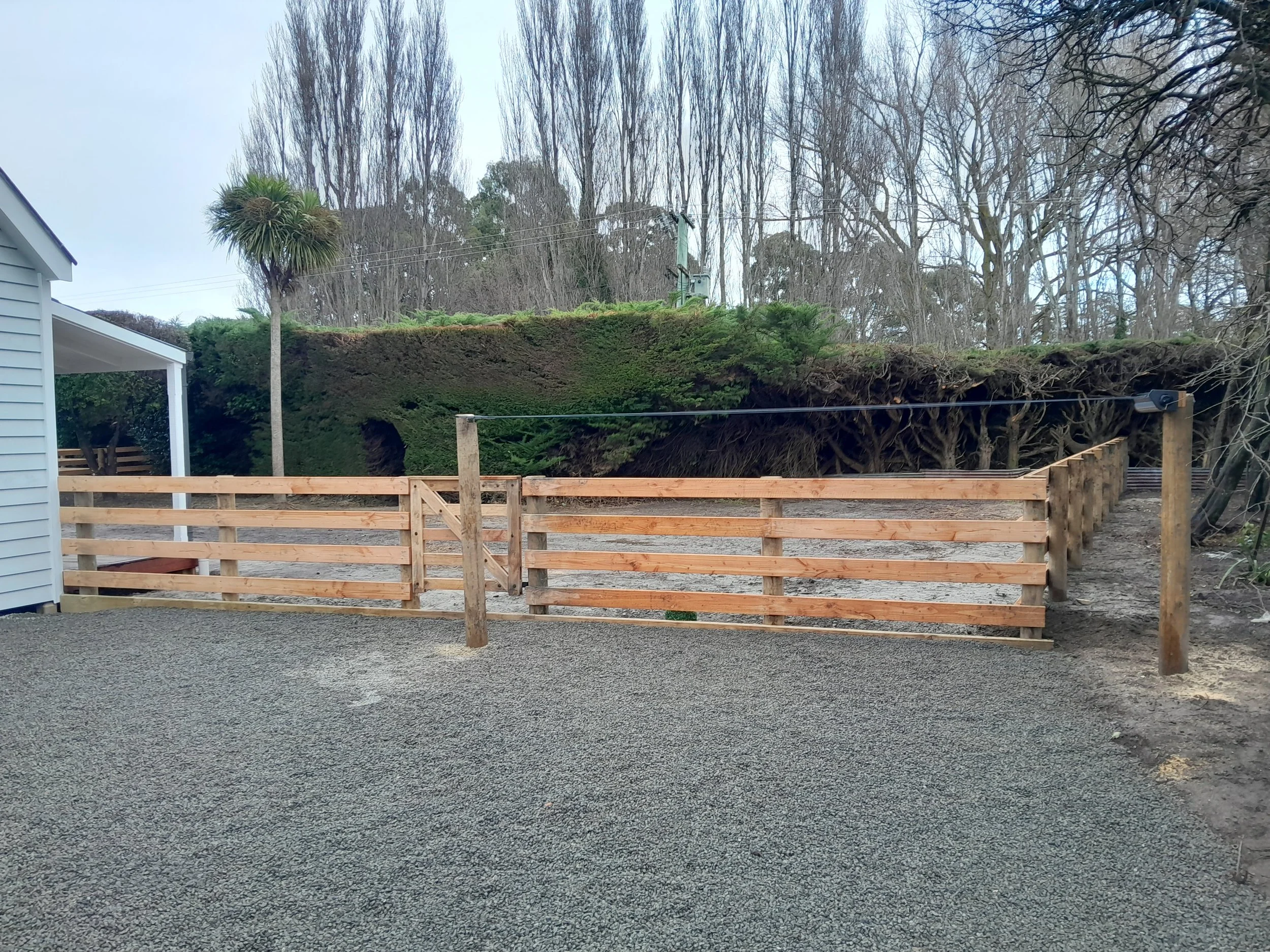 A newly constructed wooden fence with a gate, surrounding a gravel area, with a hillside covered in green bushes and trees in the background.