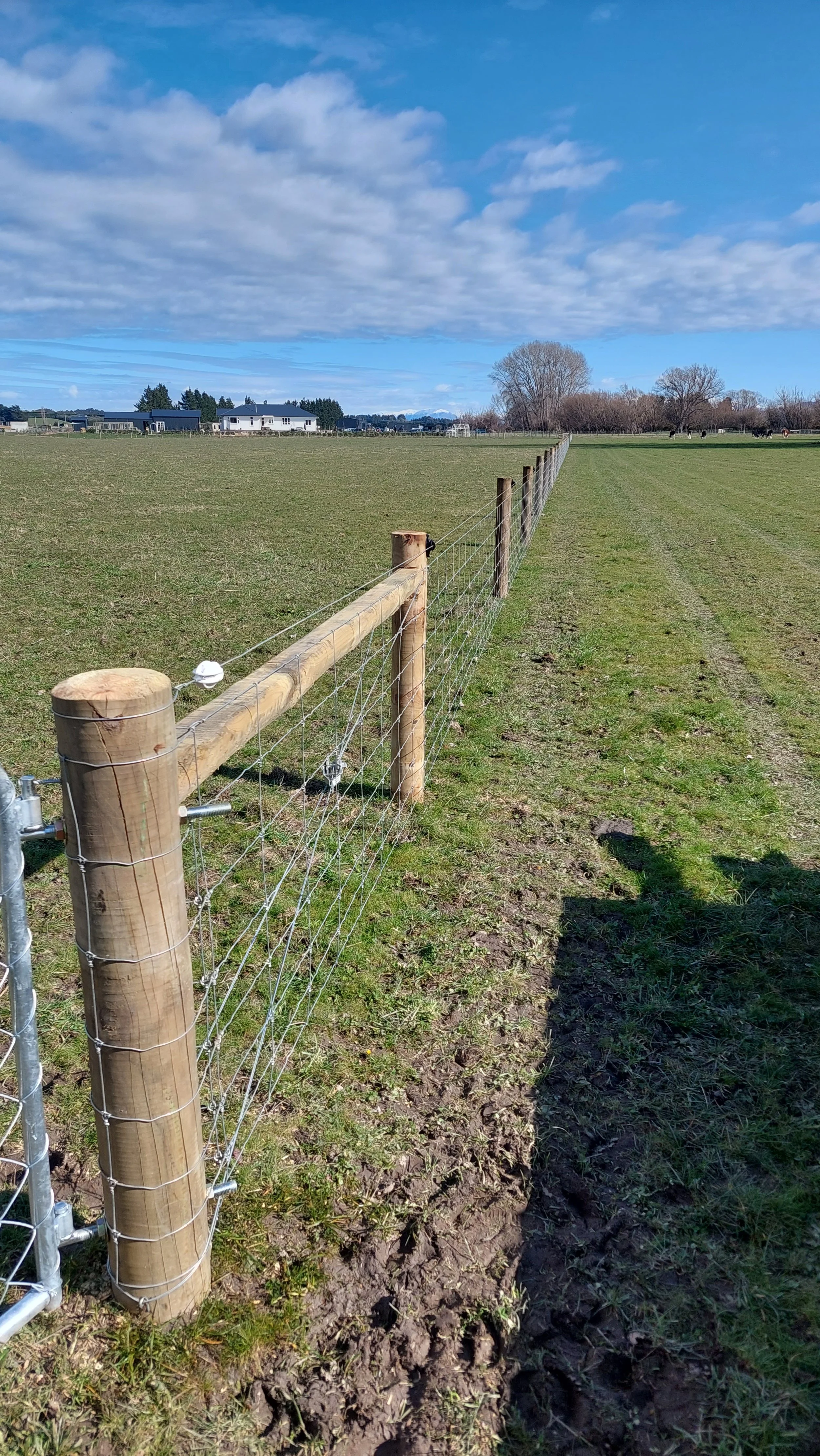 A wooden and wire fence running through a grassy field under a partly cloudy blue sky with some houses in the distance.