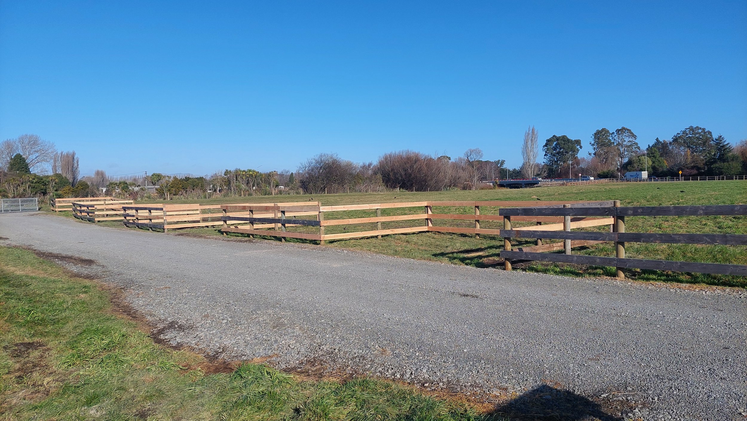 A gravel pathway running along a grassy field with a wooden fence, clear blue sky and trees in the background.
