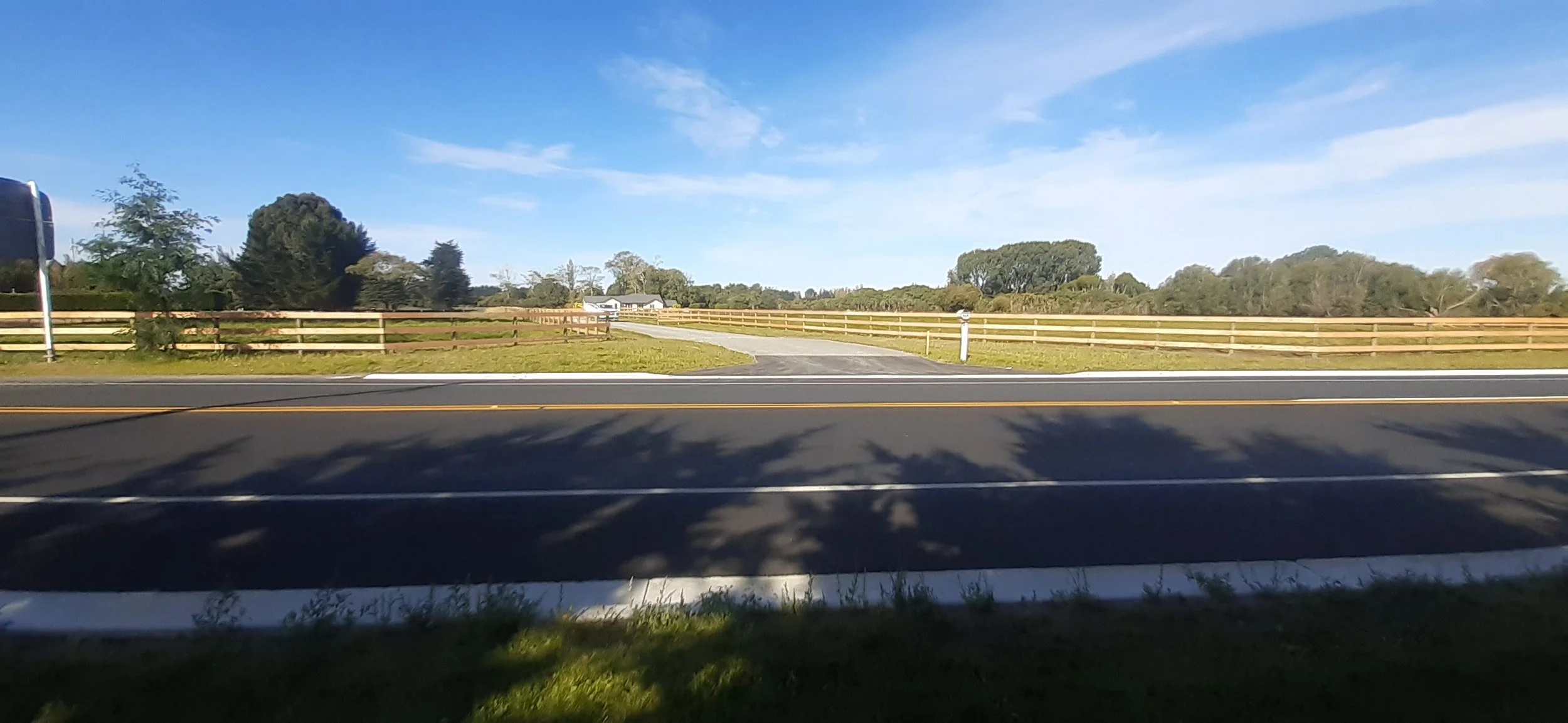 A rural road intersects with a gravel driveway leading to a farmhouse, fenced fields, trees, blue sky with some clouds.