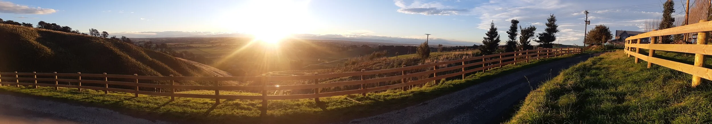 Sunset over rolling countryside with gravel road, wooden fences, trees, and houses amid grassy hills.