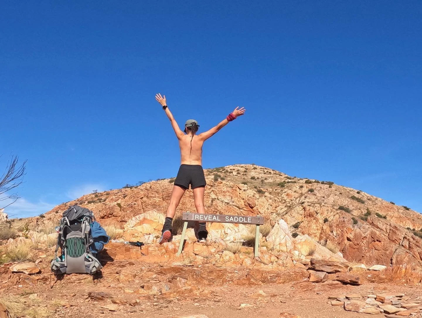 A person stands on a rocky trail with arms raised, facing a mountain, wearing black shorts and a cap, next to a backpack, under a clear blue sky.