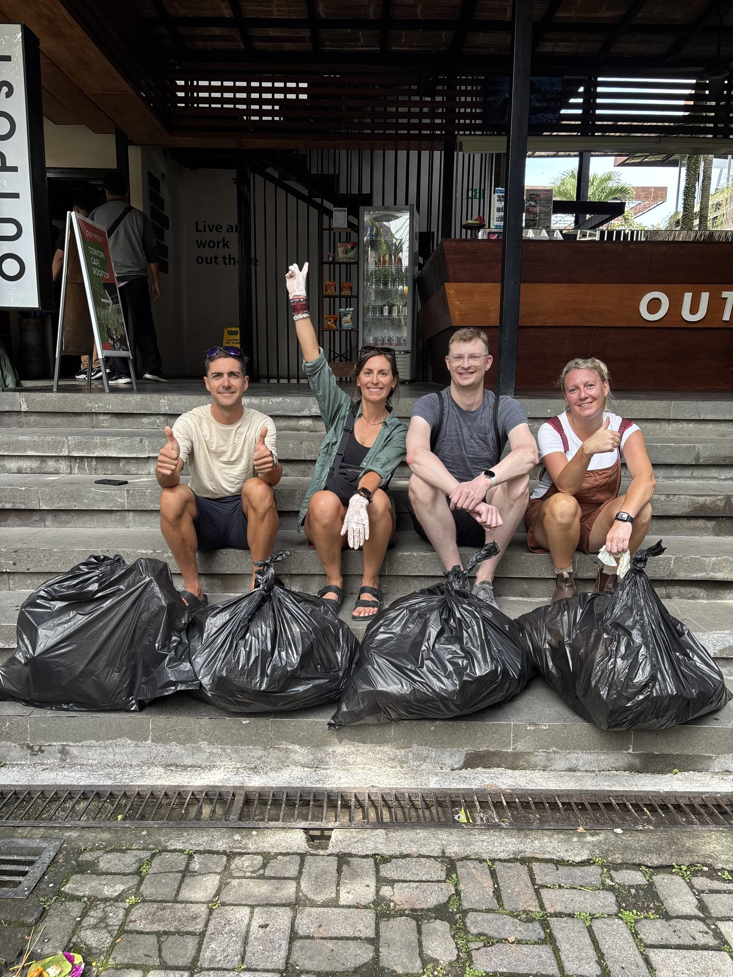 Four people sitting on steps with large black garbage bags in front of them, smiling and giving thumbs up after a cleanup event.