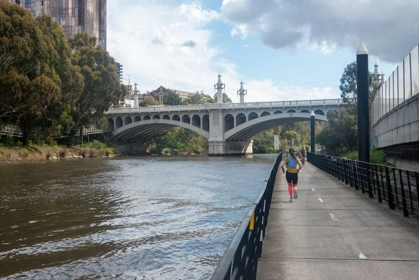 People running on a riverside pathway with a large white bridge in the background and trees on either side.