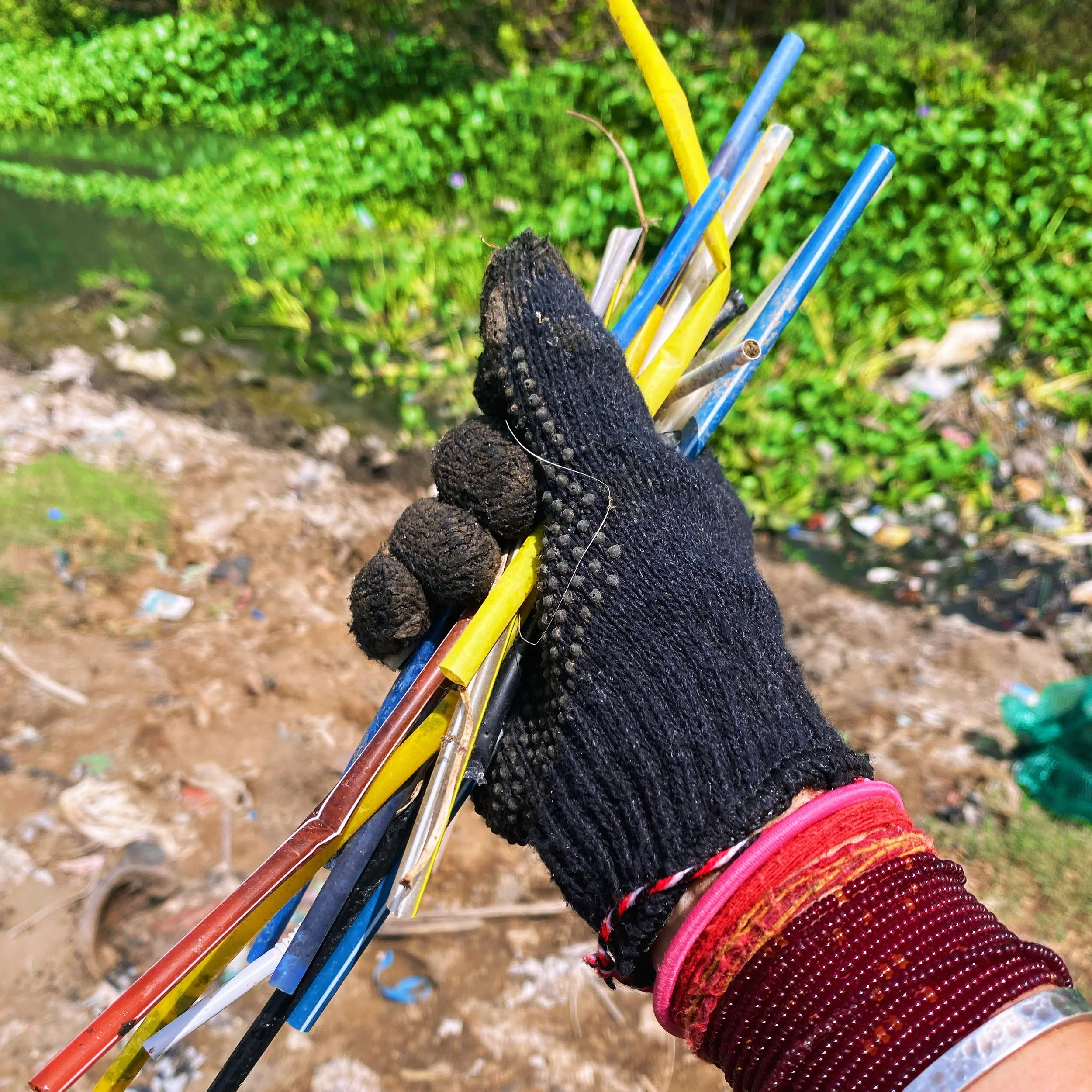 A hand wearing a black glove holding multicolored plastic straws above a dirt ground and green plants.