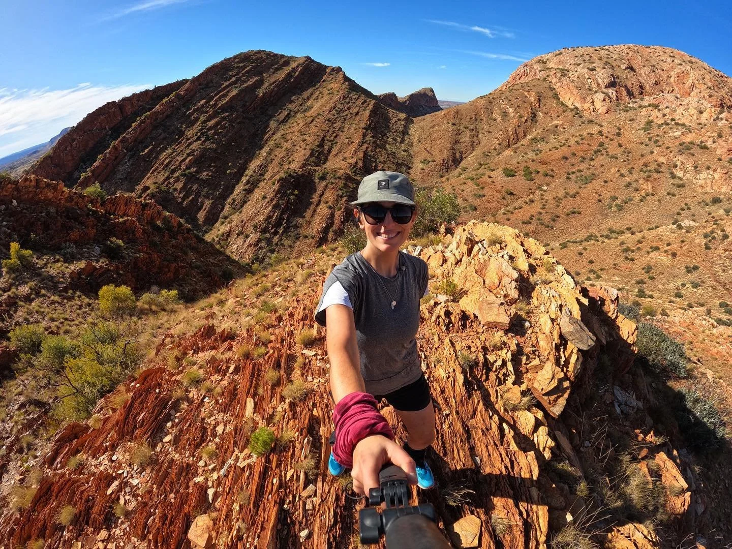 Woman in outdoor gear taking a selfie on a rocky desert mountain trail with red rock formations and sparse vegetation under a blue sky.