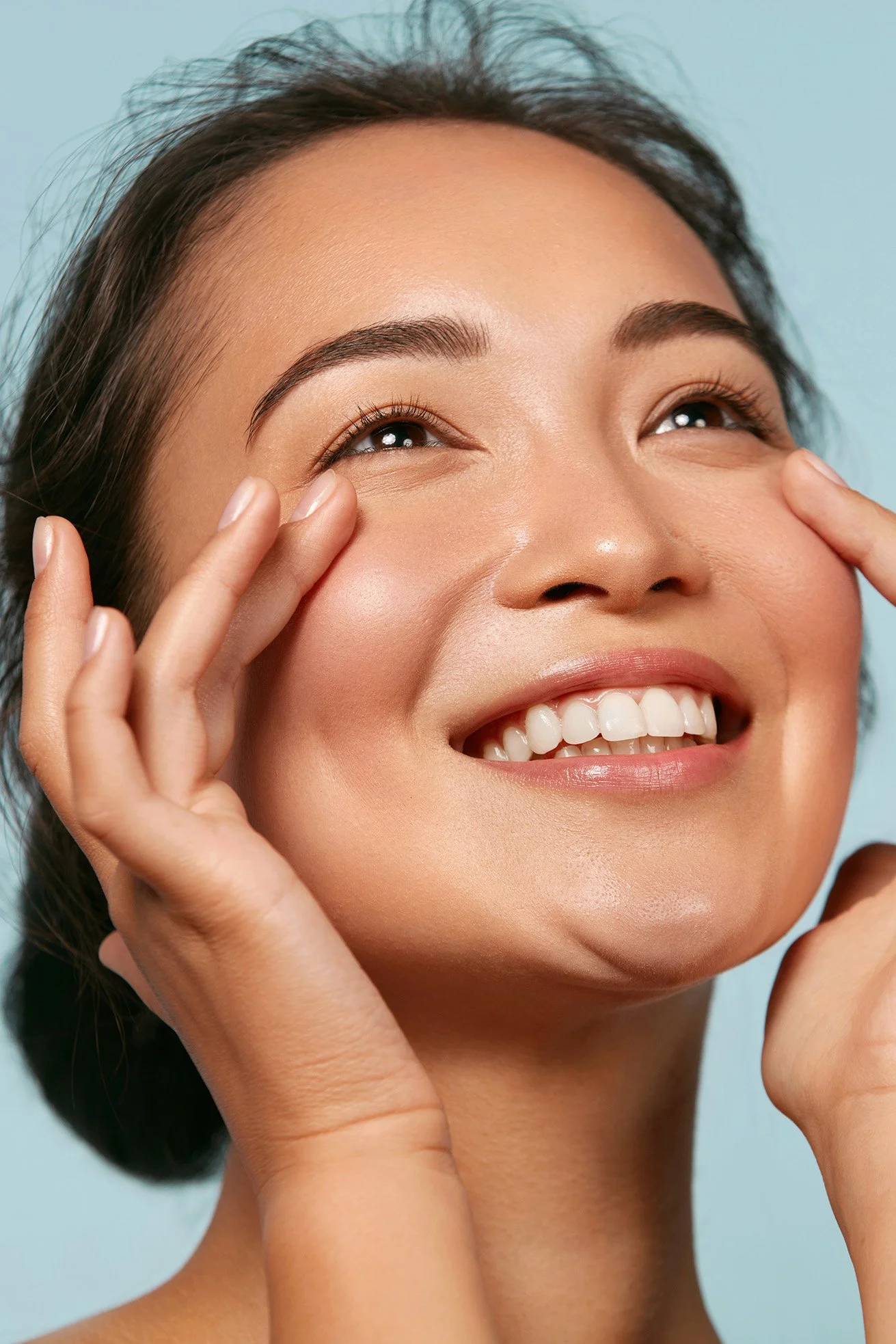 Close-up of a smiling woman with clear skin, dark eyebrows, and dark hair, touching her face and looking upward against a light blue background.