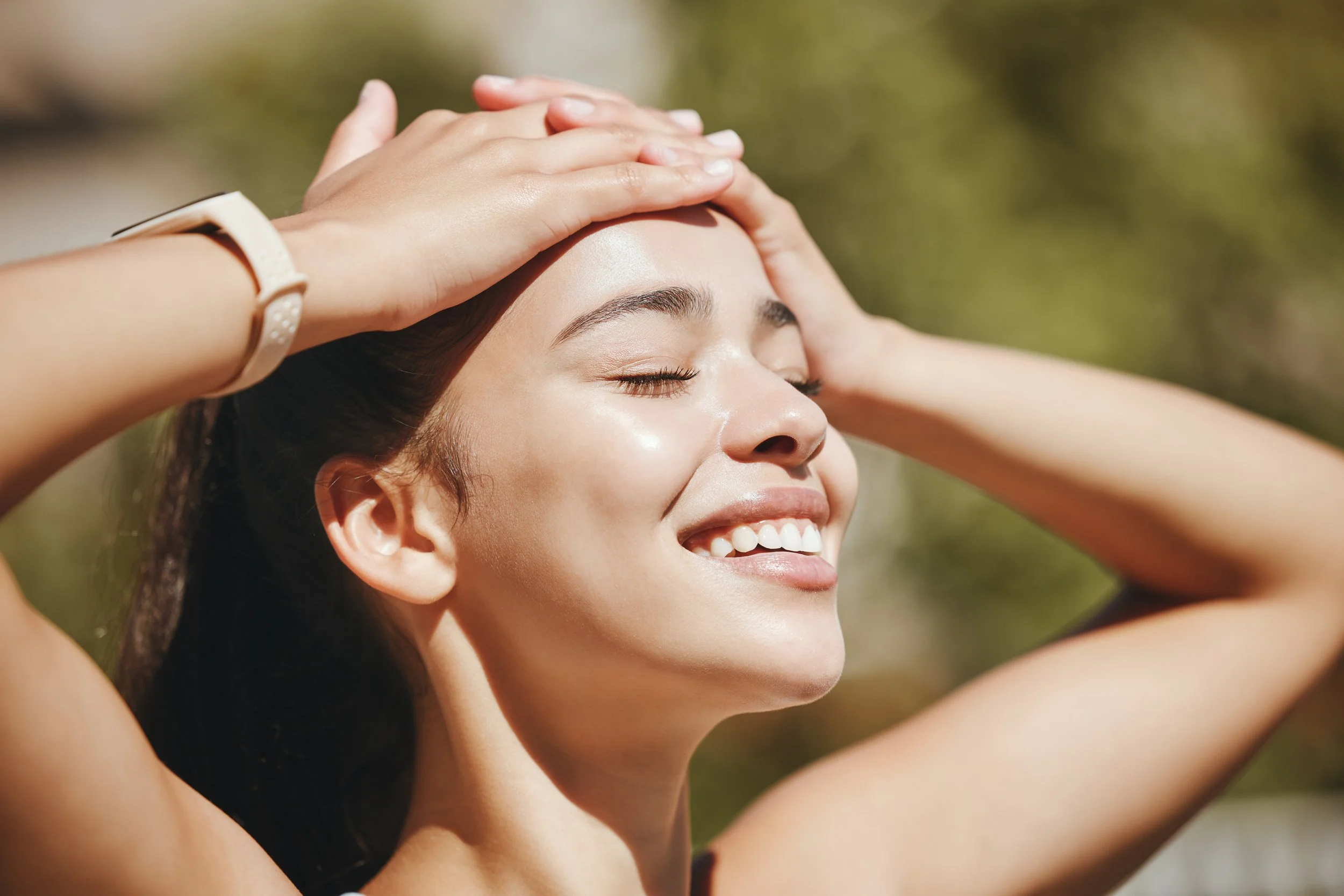 A woman smiling with her eyes closed, holding her head with her hands outdoors in the sunlight.
