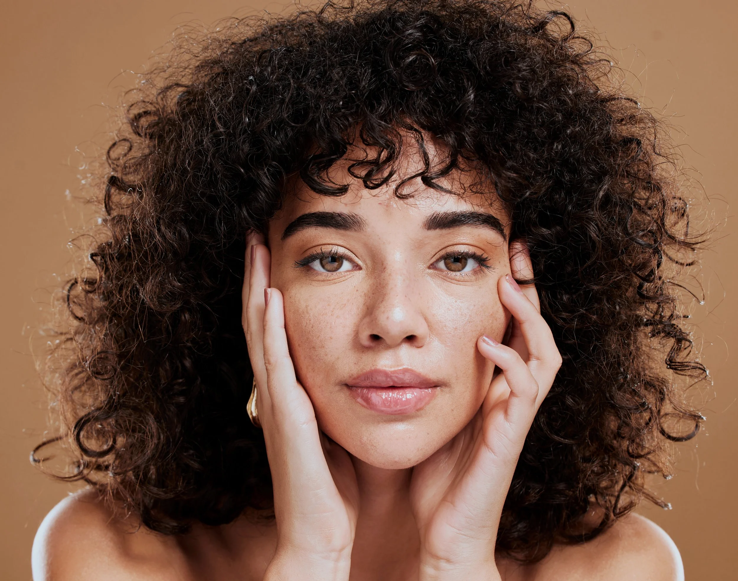 Close-up of a young woman with curly dark hair and light brown eyes, holding her face with her hands against a beige background.