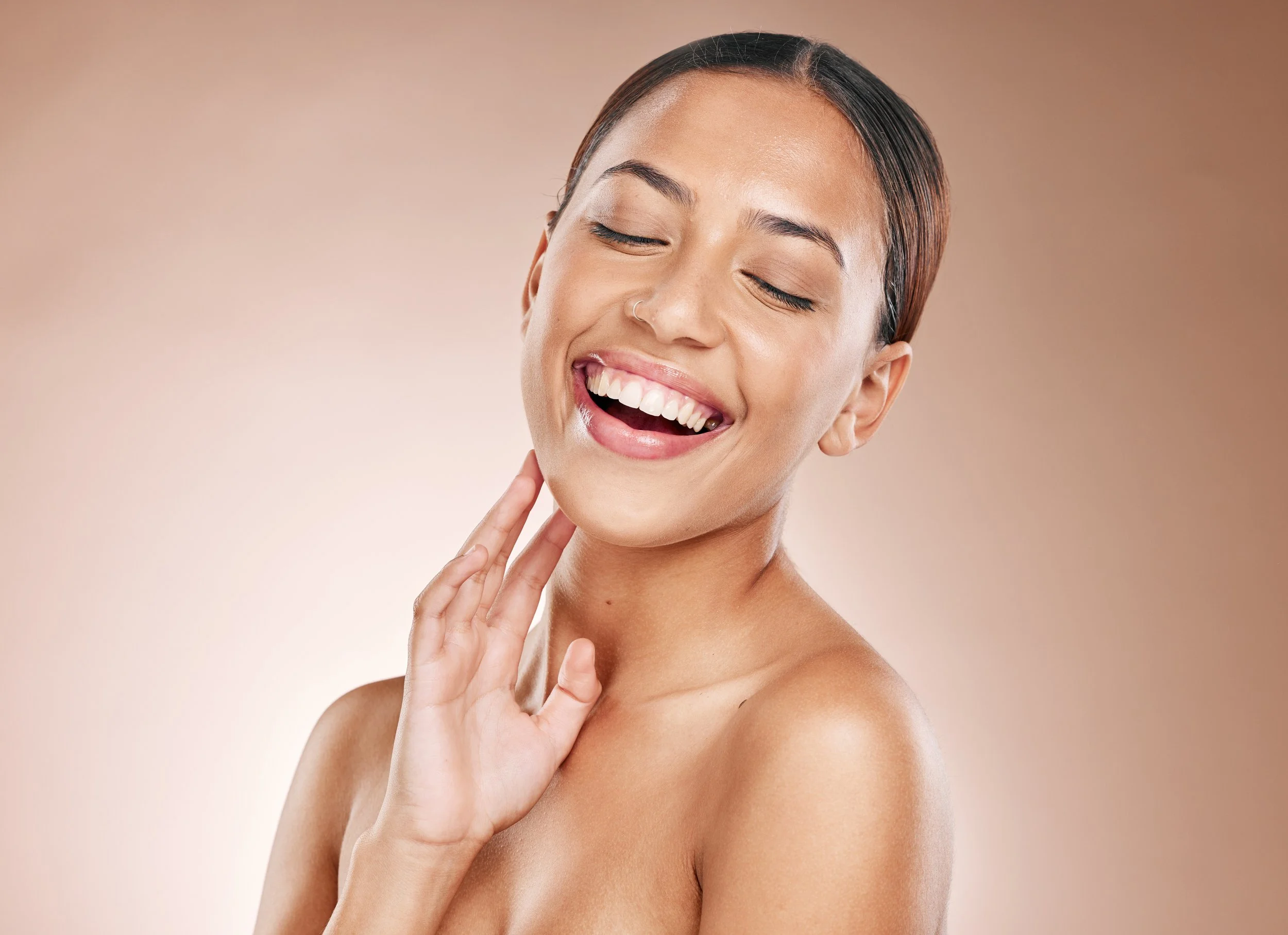 A young woman with short dark hair and a nose piercing smiling with her eyes closed, touching her neck with her hand, against a neutral background.