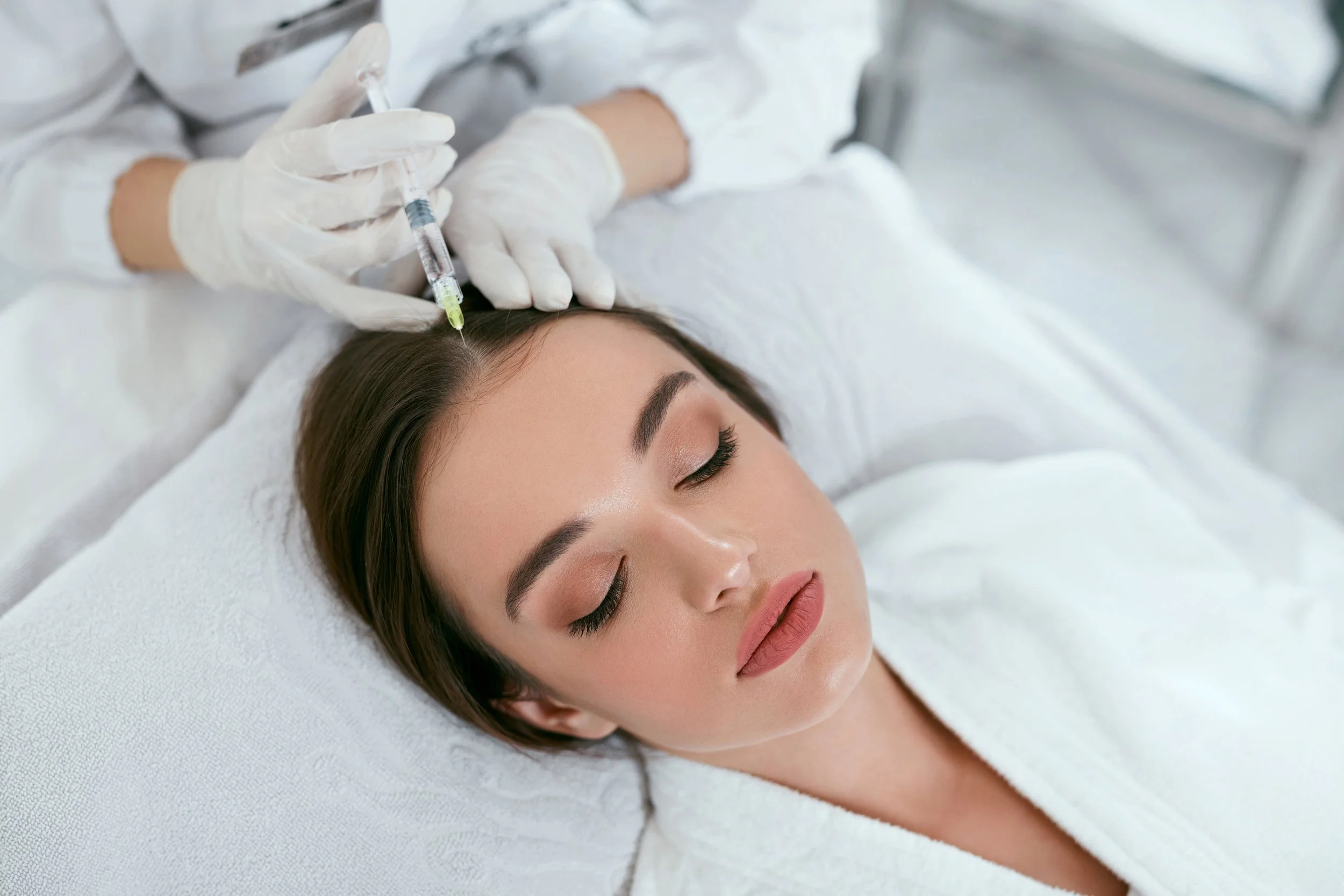 A woman receiving a forehead injection from a healthcare professional wearing white gloves, while lying on a bed with white sheets in a medical setting.