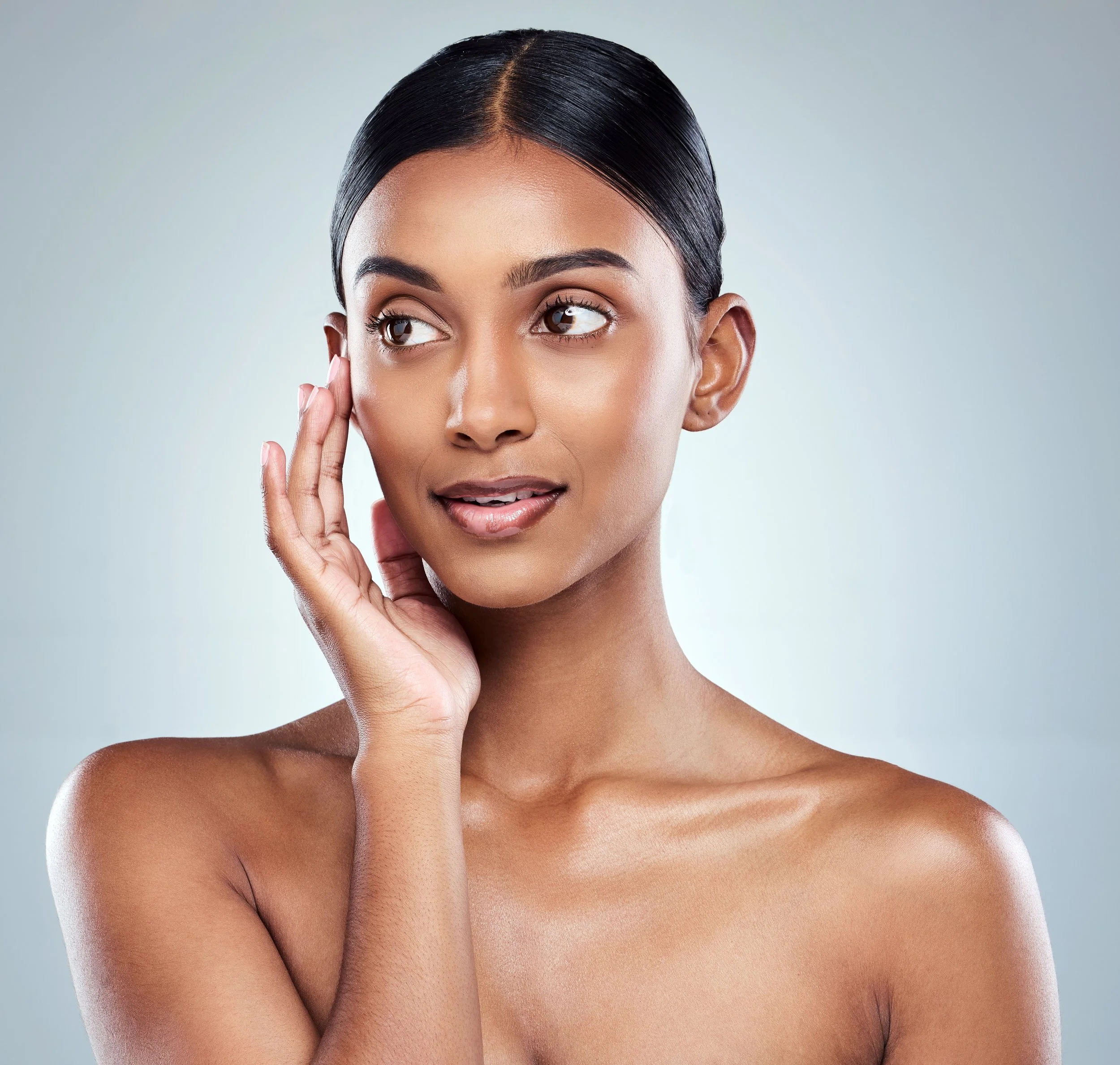 A young woman touching her face with her right hand, looking to the side, with smooth skin and dark hair pulled back.