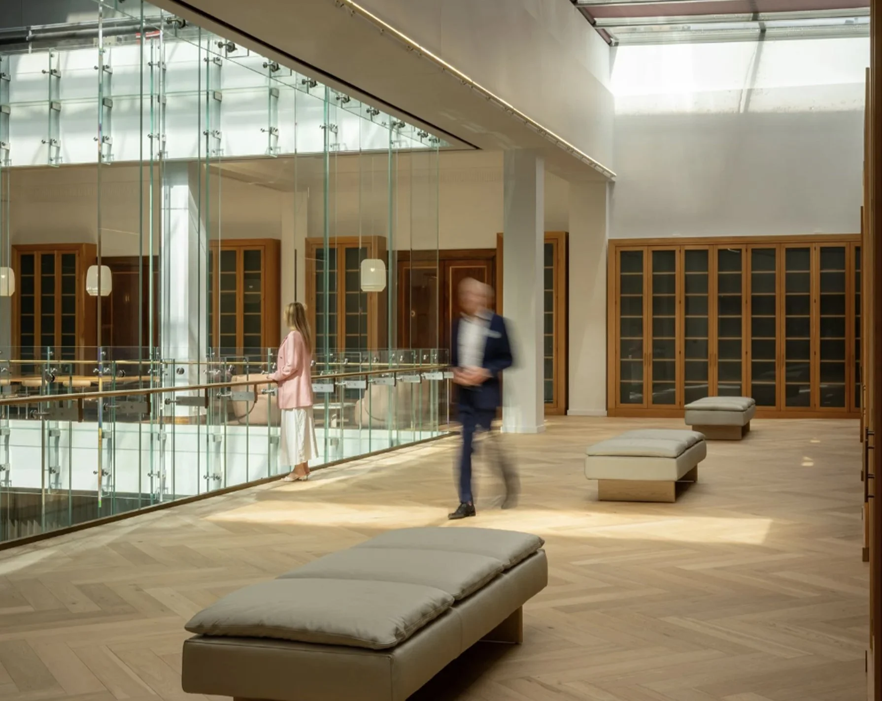 Modern interior space with wooden floors, glass railing, and wooden cabinets. Two cushioned benches are in the foreground, with three people walking and standing in the background. Natural light comes from the large ceiling window.