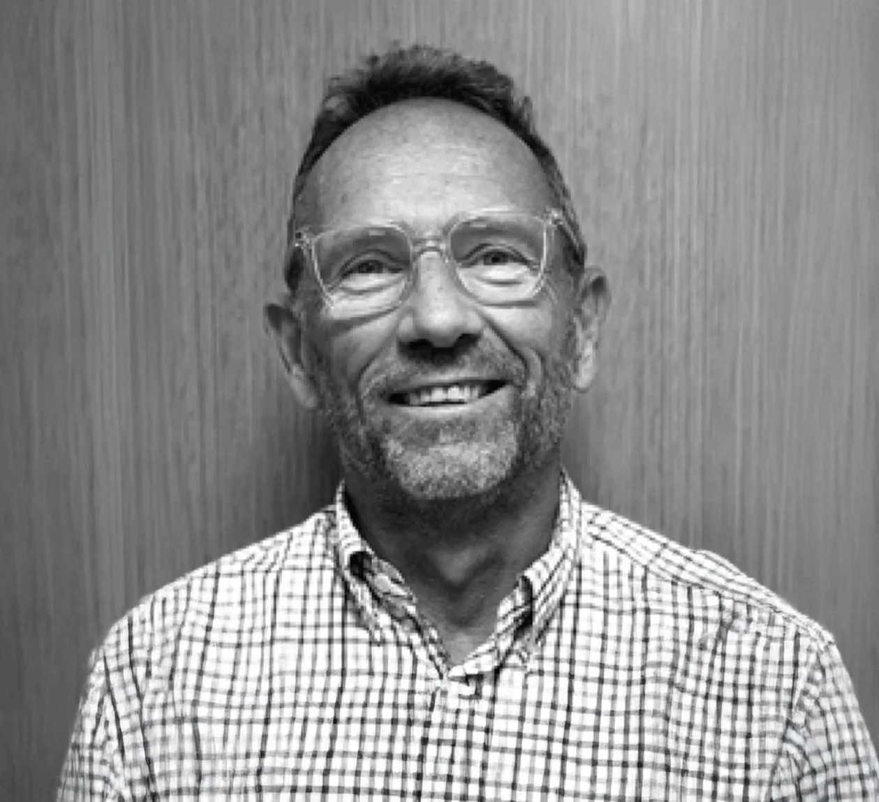 A smiling middle-aged man with glasses and a beard, wearing a checkered shirt, against a wooden wall background.