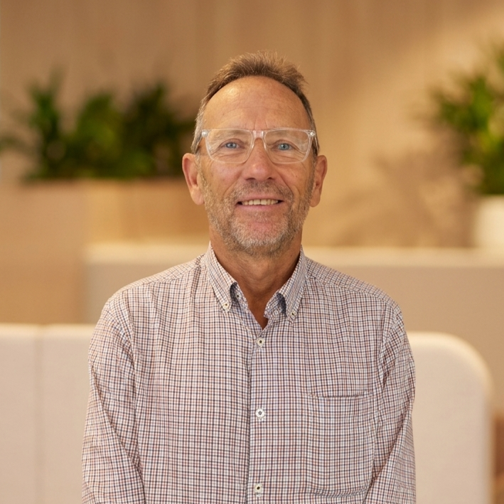 A middle-aged man with glasses and a beard, smiling, in an indoor setting with plants in the background.