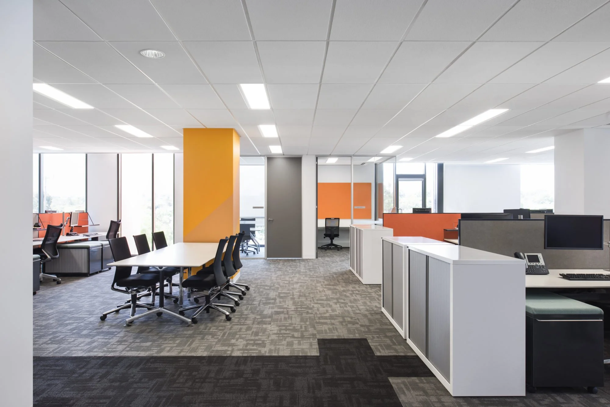 Modern office space with open layout, white desks, black office chairs, orange accents on walls, large windows, and ceiling with recessed lighting