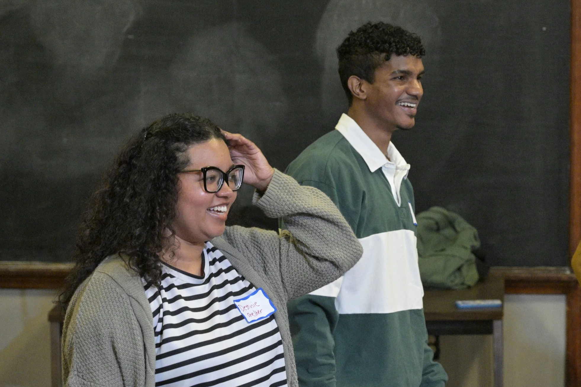 A woman with curly dark hair, glasses, and a beige sweater, smiling and touching her head, standing next to a man with short curly dark hair, smiling, wearing a green and white rugby shirt, inside a room with a chalkboard background.