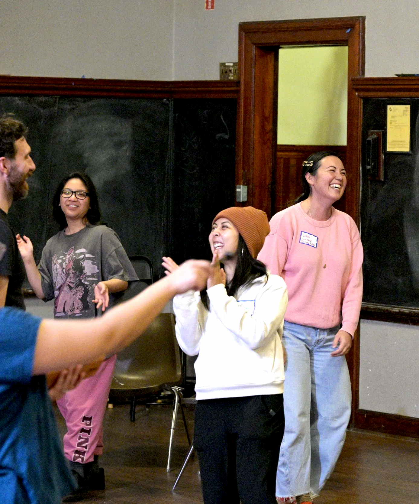 A group of people standing and chatting in a room, smiling and laughing. Four women and one man are visible, with some wearing casual clothes and one woman wearing a brown beanie.