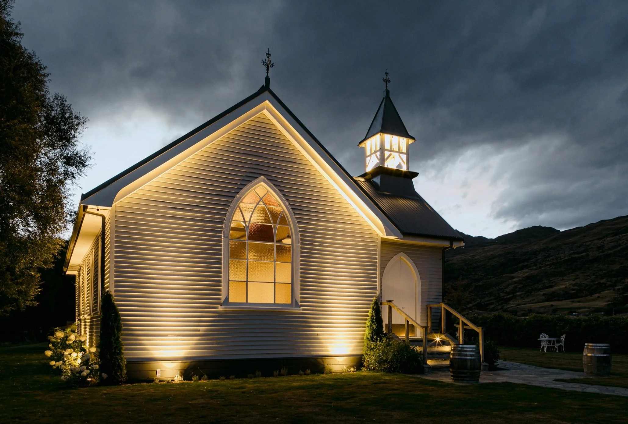 A small church with illuminated exterior lighting, white siding, pointed arch stained glass window, set against a dark, cloudy sky, with a mountain landscape in the background.