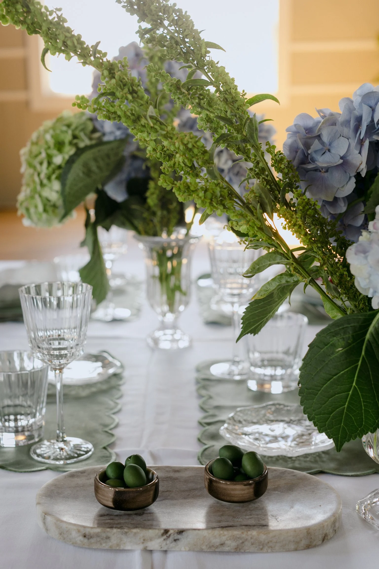 Elegant dining table with floral centerpiece, green olives in small bowls, crystal glasses, and place settings, illuminated by soft natural light.
