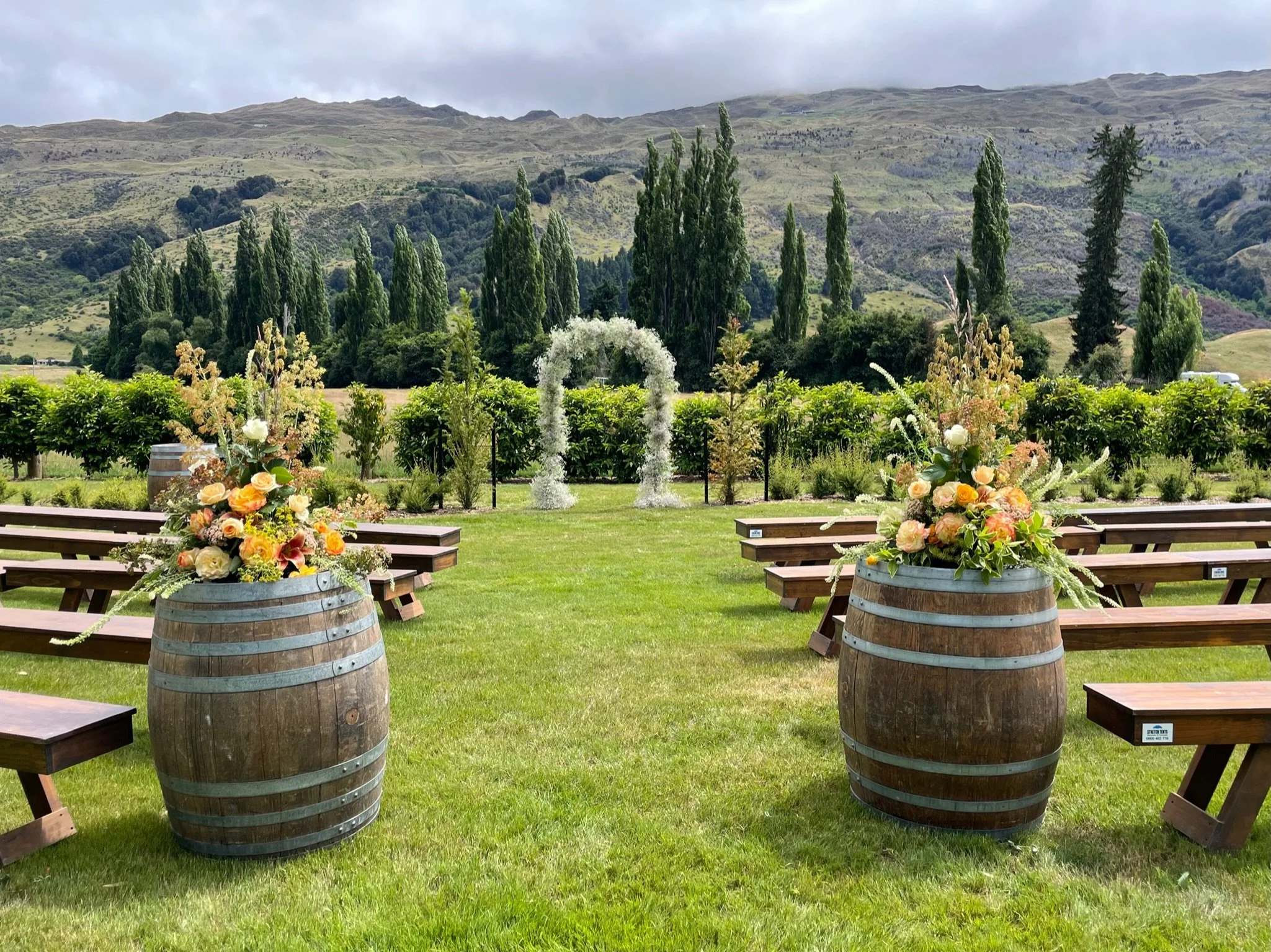 Outdoor wedding ceremony setup with wooden benches, floral arrangements in wooden barrels, and a floral arch in a scenic mountain landscape