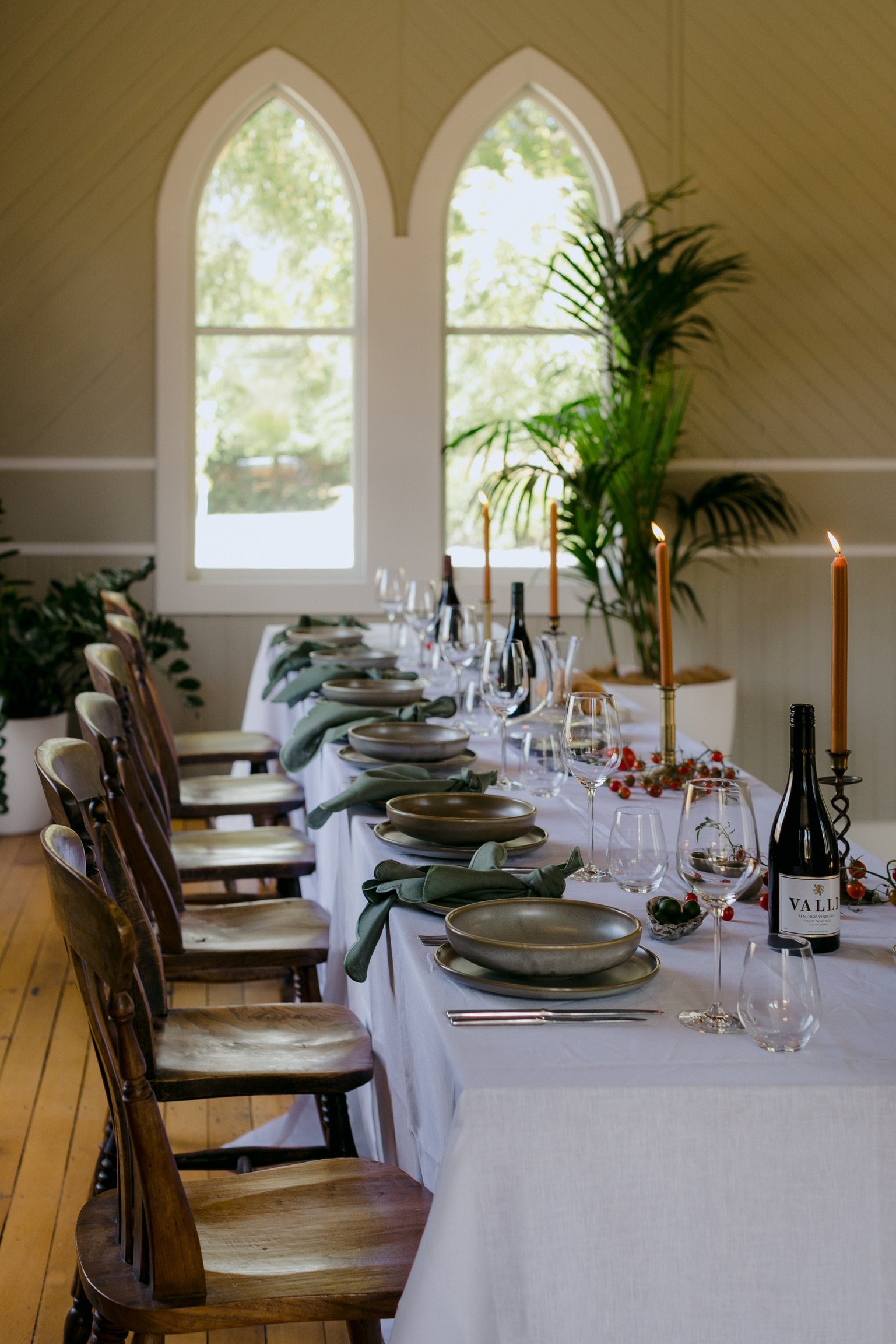 A table set for a formal dinner with white tablecloth, green napkins, wine glasses, and plates, decorated with candles and cherry tomatoes in a room with windows and wooden floors.